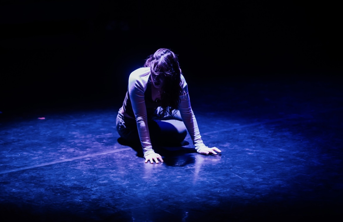 A solo dancer kneeling on the floor under a blue spotlight. Her hair is obscuring her face. But she looks distraught and disheveled. She's resting heavily on her hands.