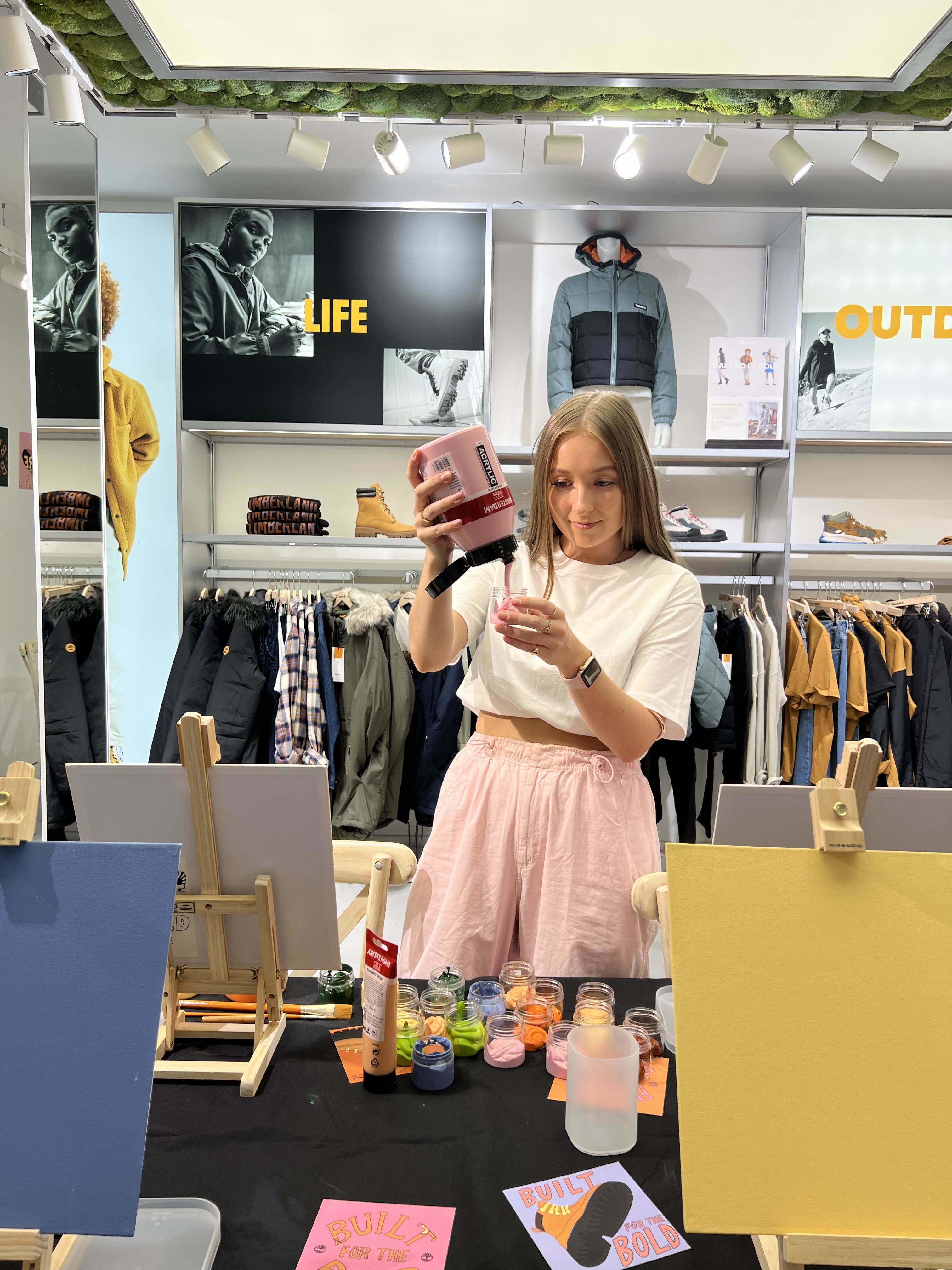 Tegan Price hosting a workshop for Timberland, pouring pink paint into a glass jar with with paint jars and canvases in front, clothes behind.
