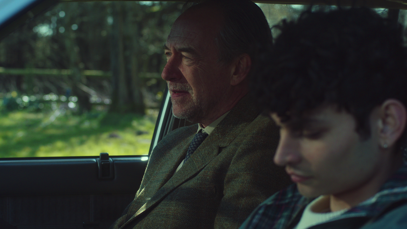 Two men inside a car, older man driving in olive jacket, younger man as passenger, green foliage visible through windows.