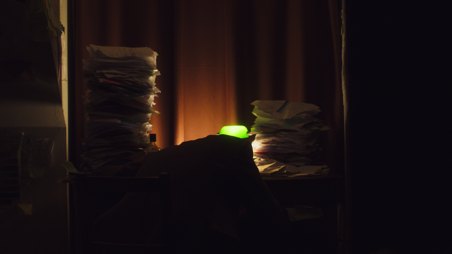 Dark interior with stacks of books or papers on surfaces, illuminated by warm orange light and a bright green glow from small object.