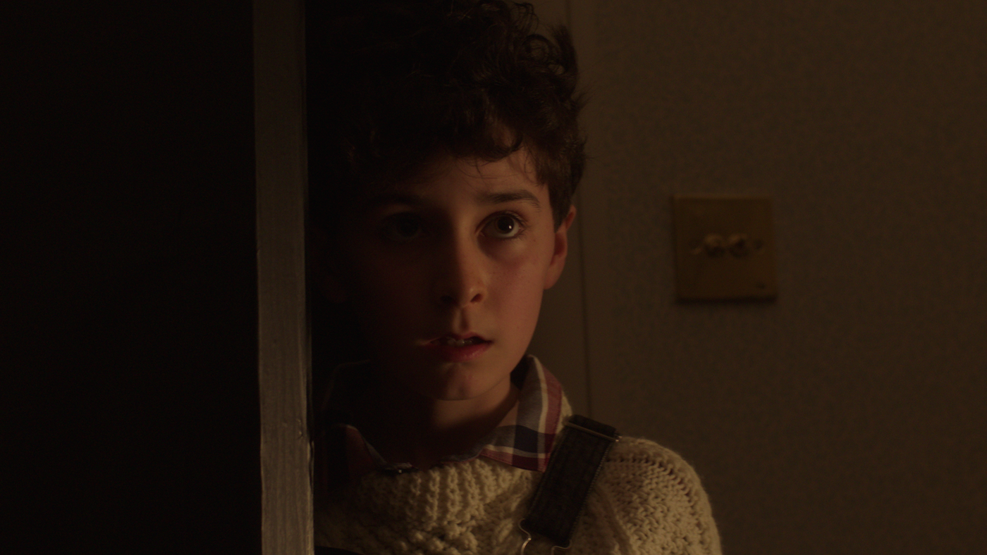 Young man with curly dark hair wearing cream-coloured jumper, lit by warm orange light against dark background.