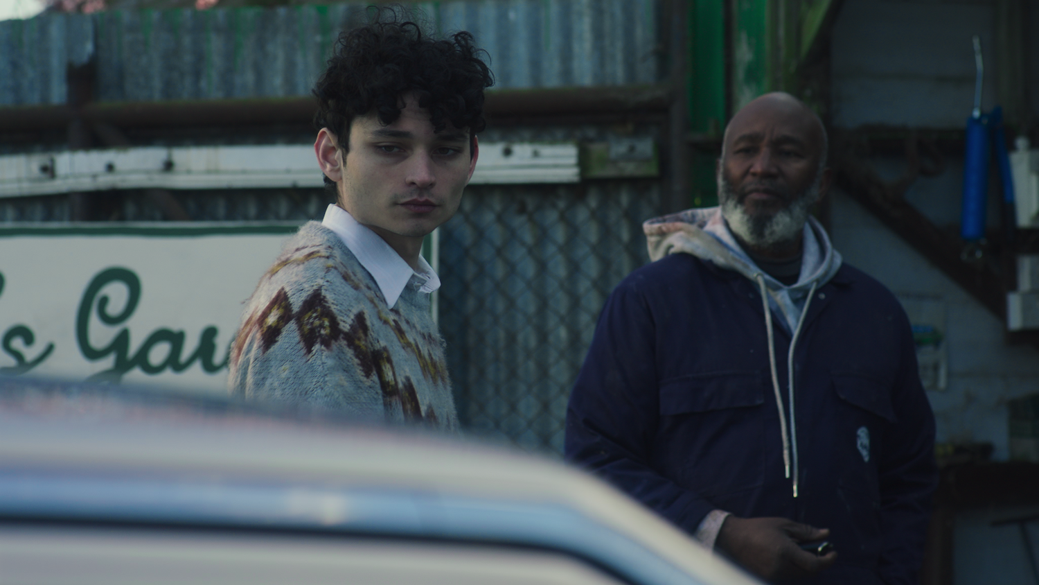 Young man with curly hair in patterned jacket and older man in dark hoodie standing outside shop, viewed through car window.