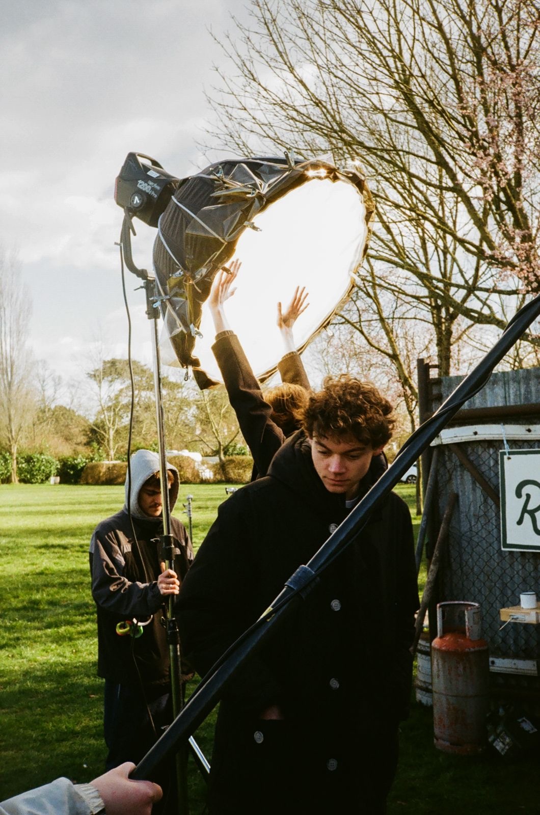 Film crew with lighting equipment and reflector disc in park setting, bare trees and van visible in background.