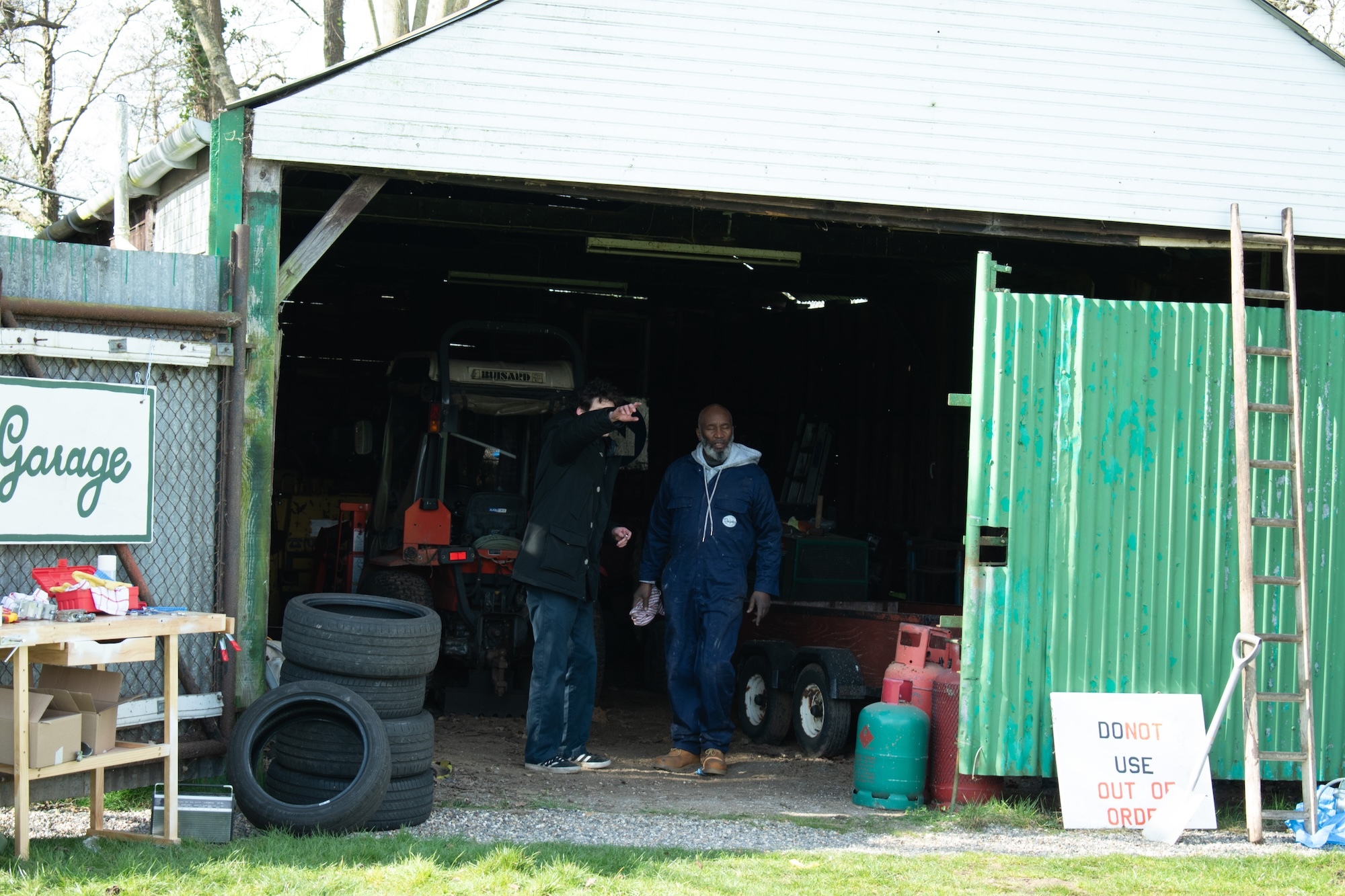 Open garage with green corrugated metal siding, white roof, two people inside, tyres scattered outside, "Garage" sign on left fence.