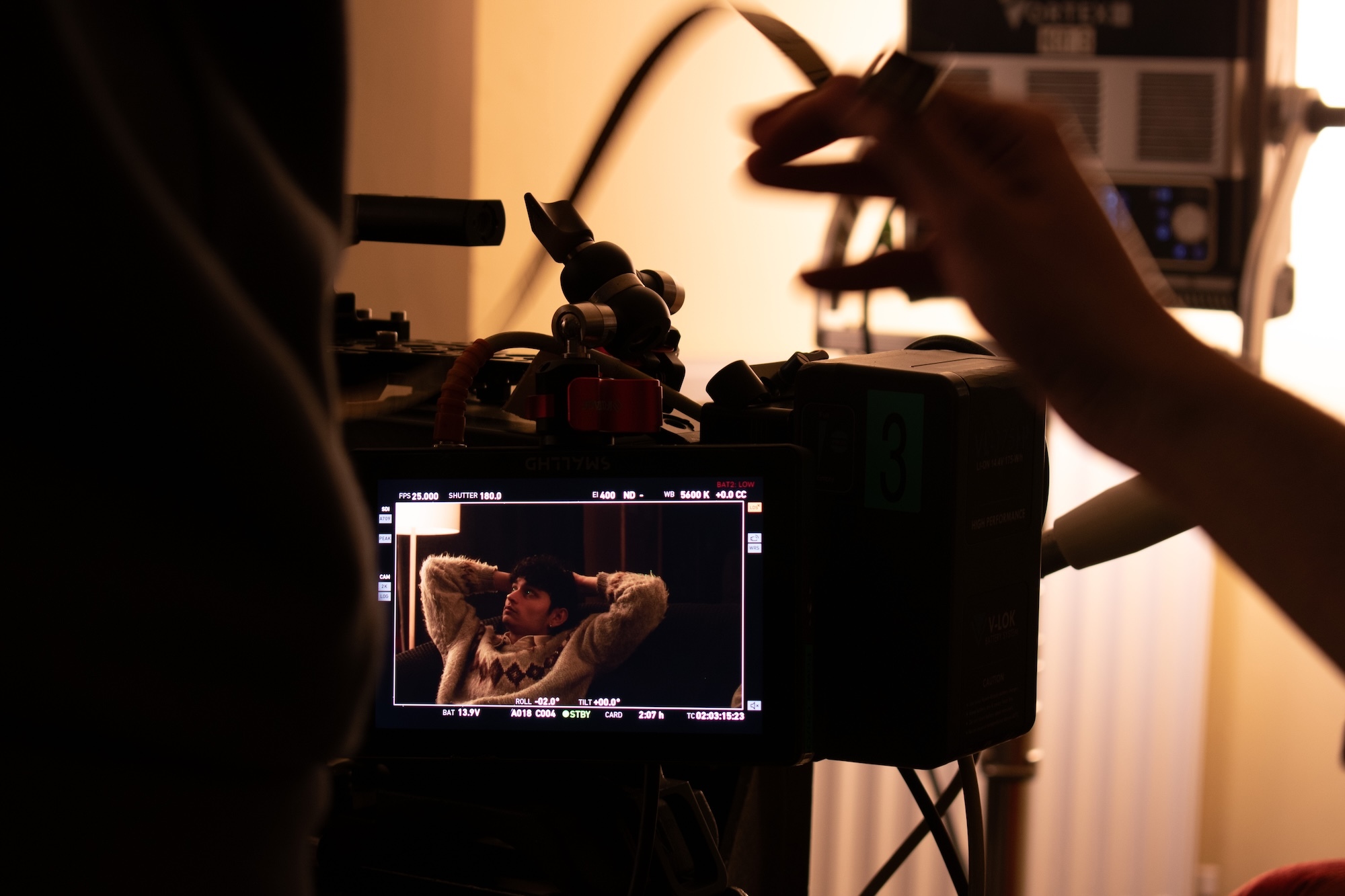 Hands operating professional camera with LCD screen displaying tattooed subject, warm lighting, indoor filming setup.