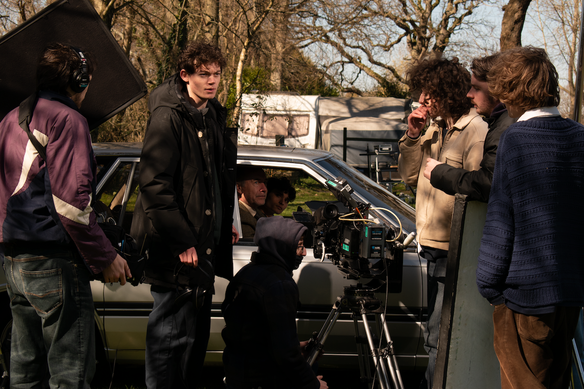 Film crew shooting scene with actors around car, camera equipment visible, bare trees in background, outdoor location.