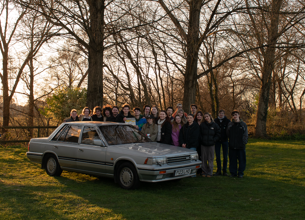 Large group of people standing behind grey saloon car on grass field with bare trees in background during golden hour light.