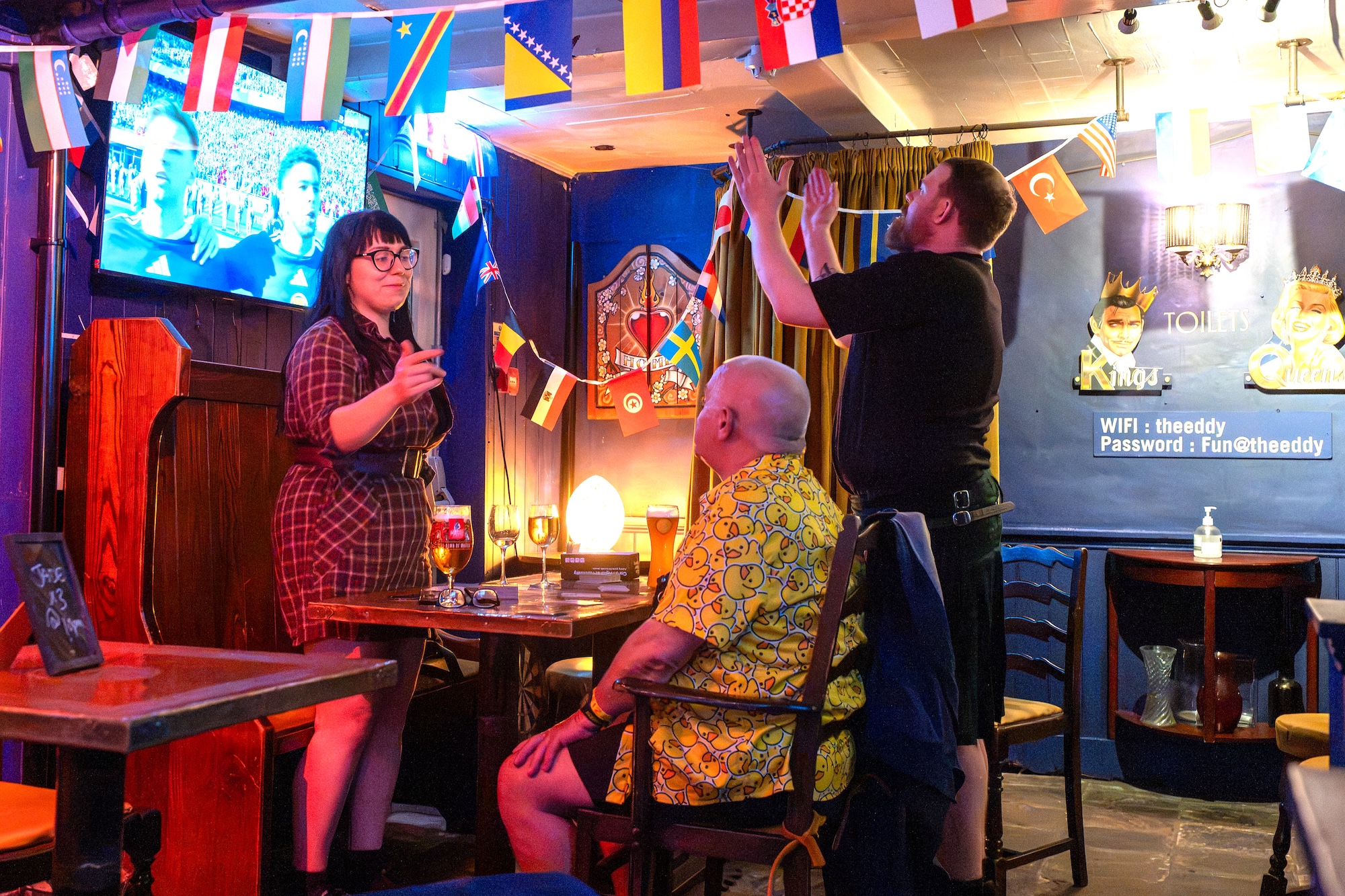 Three people in a pub decorated with international flags, with one man adjusting a hanging flag near a wall-mounted screen.
