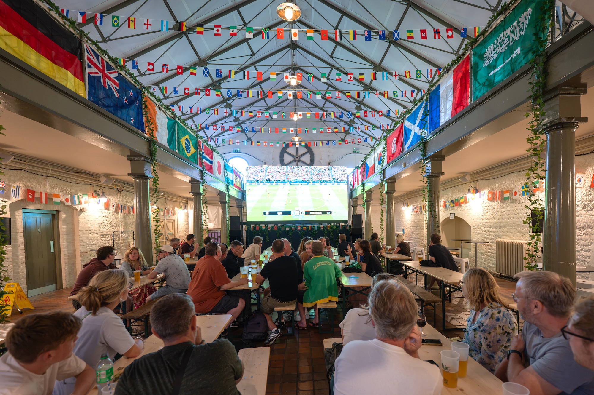 A crowd of people sits at long tables in a decorated indoor hall, with international flags hanging from the ceiling and a large screen at the far end.