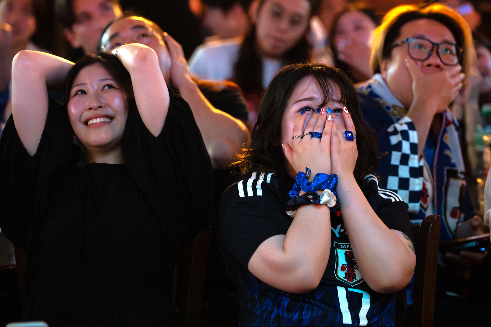 Two women in black shirts sit among a crowd, one with her hands on her head and the other covering her mouth, with other spectators blurred in the background.