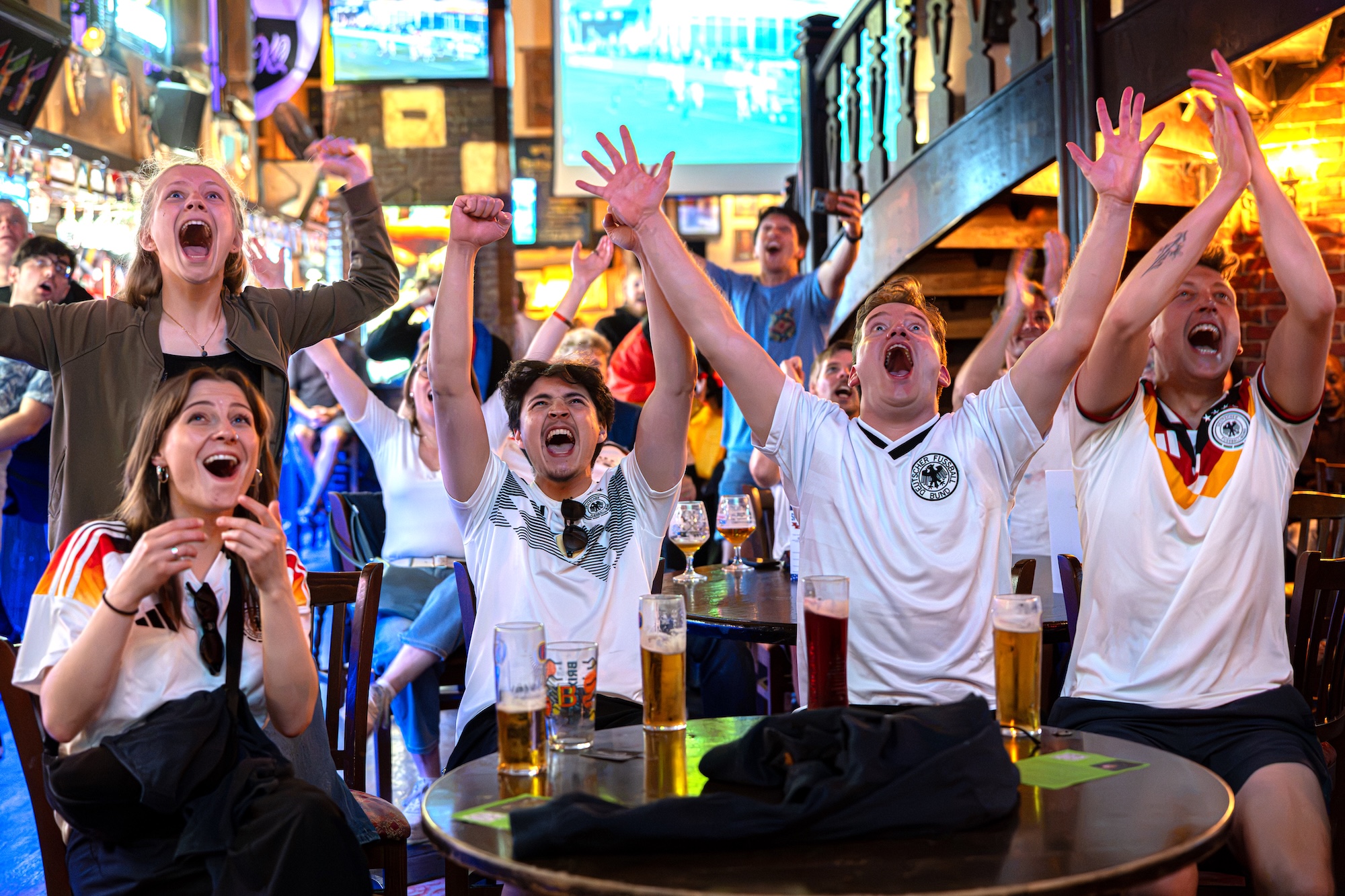 A group of football fans in white shirts raise their arms around a table with beer glasses in a busy bar with screens and stairs in the background.