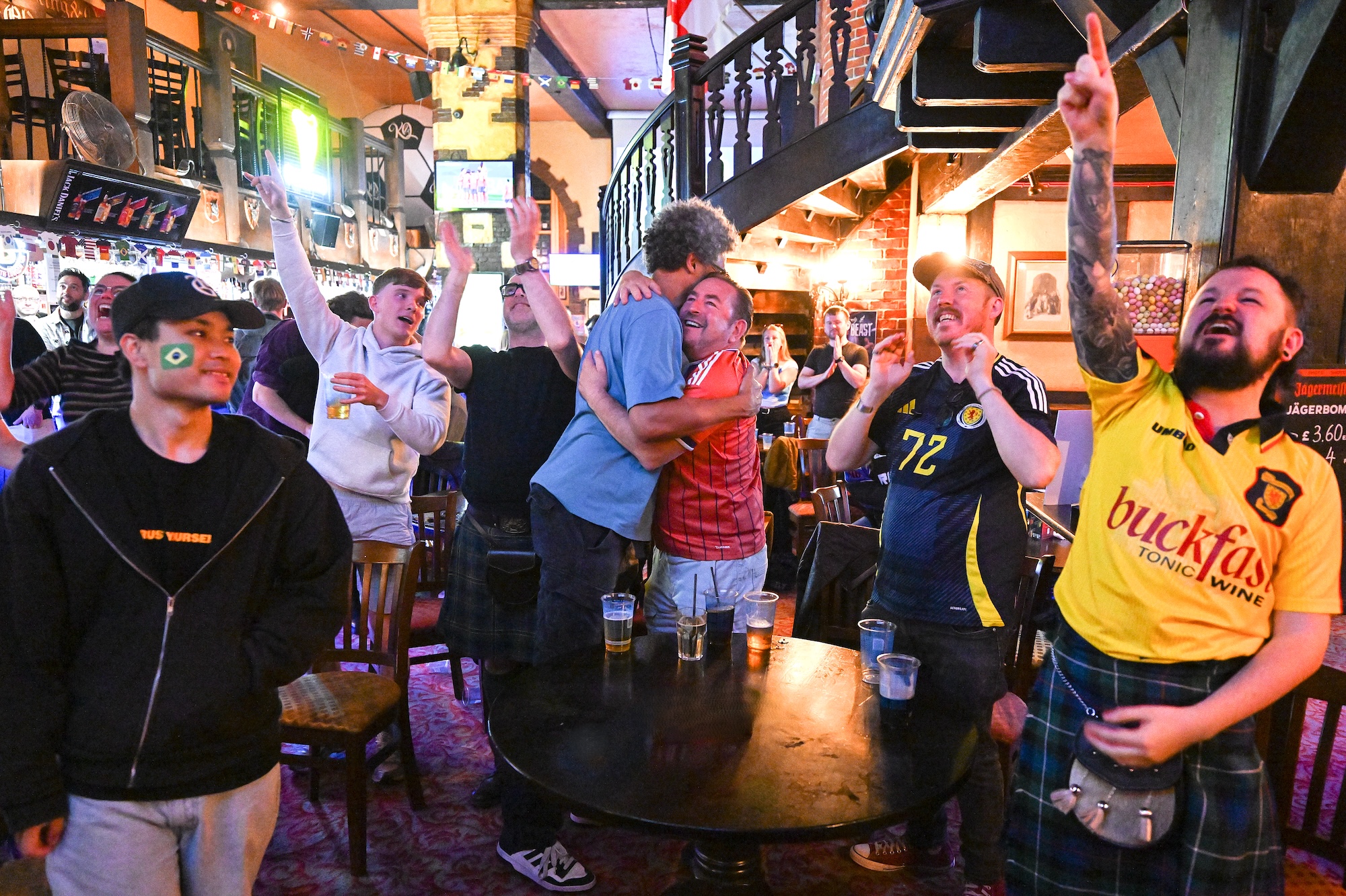Men gathered in a pub watching a sports broadcast, with one man in a yellow football shirt pointing upwards beside a staircase and tables.