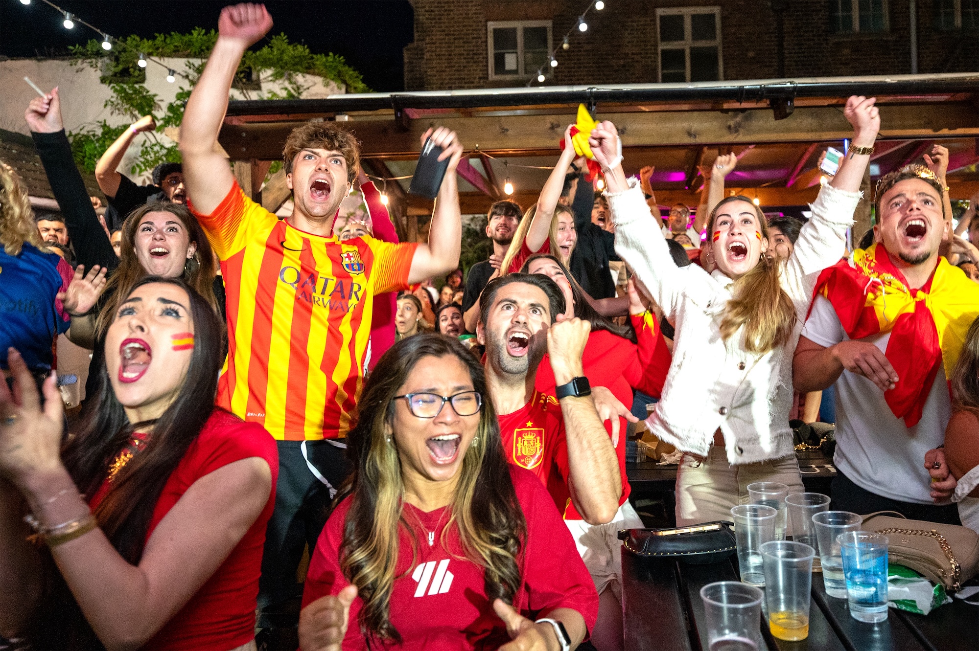 A group of football fans in red and yellow shirts cheering with raised arms at a crowded outdoor venue, with plastic cups and tables in the foreground.