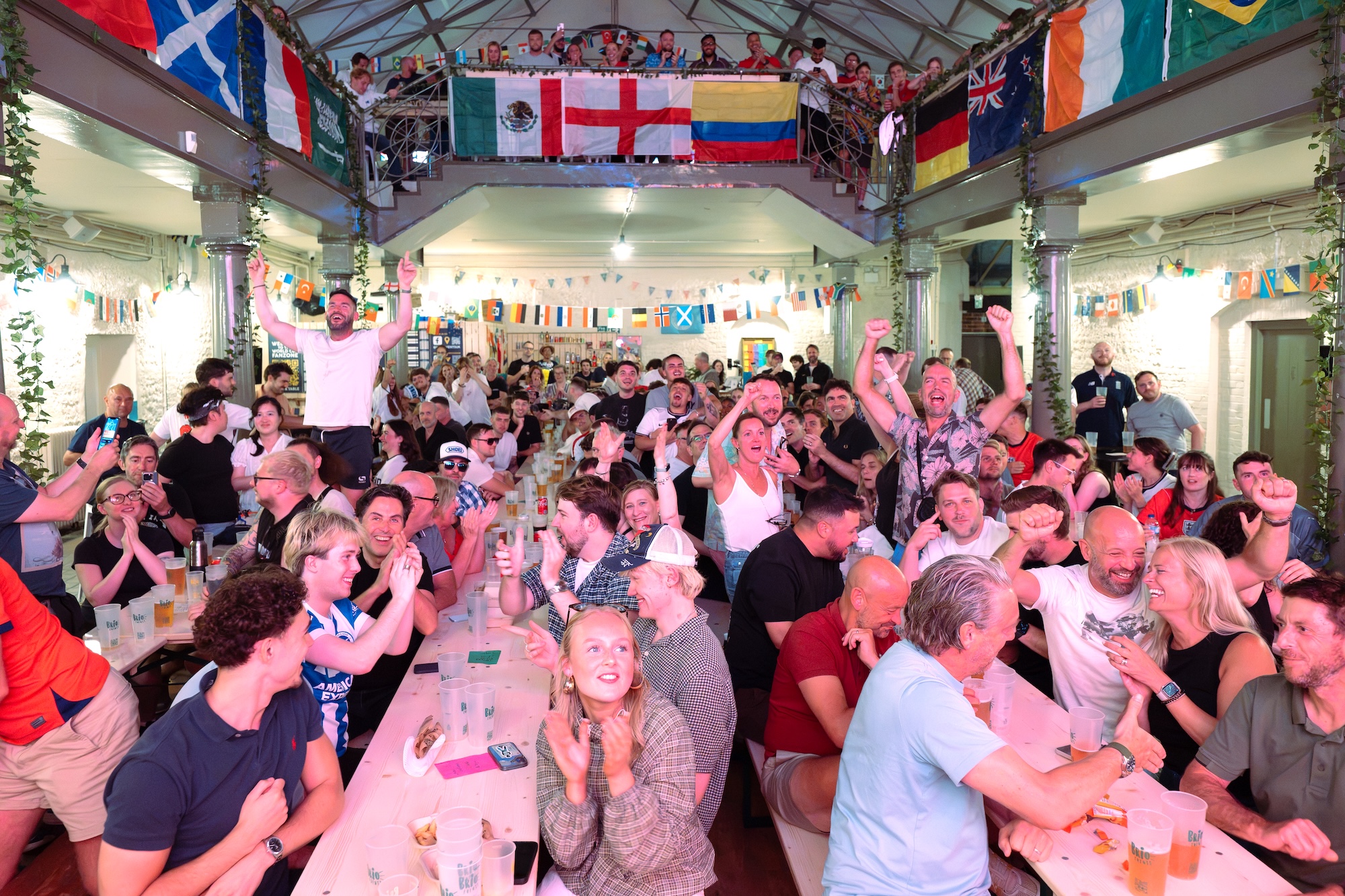 A crowded indoor bar or hall filled with people sitting at long tables and standing on a mezzanine, with national flags hanging from the upper level.