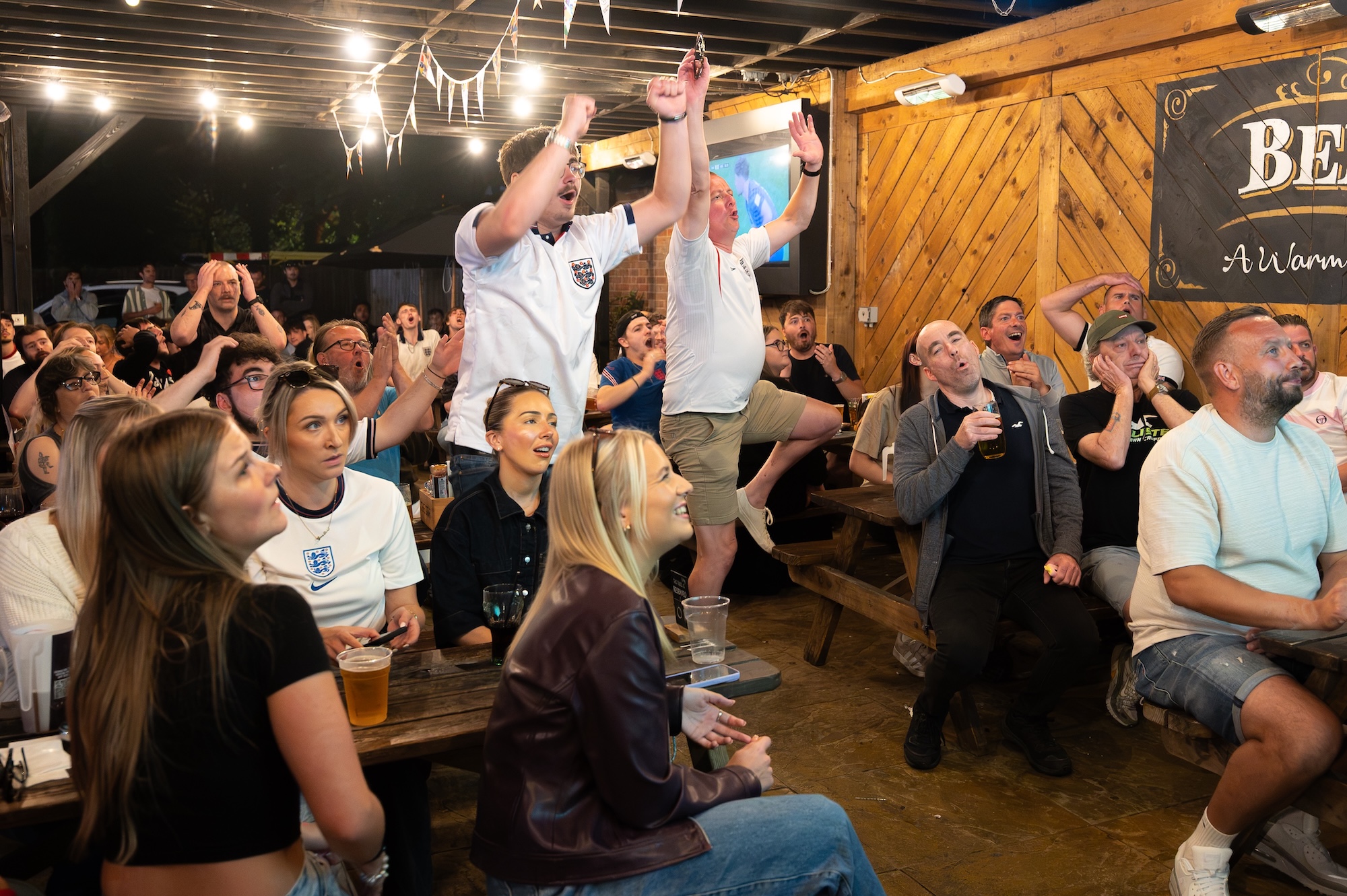 A group of people seated on wooden benches in a covered beer garden, with two men standing and raising their arms as others clap and watch.