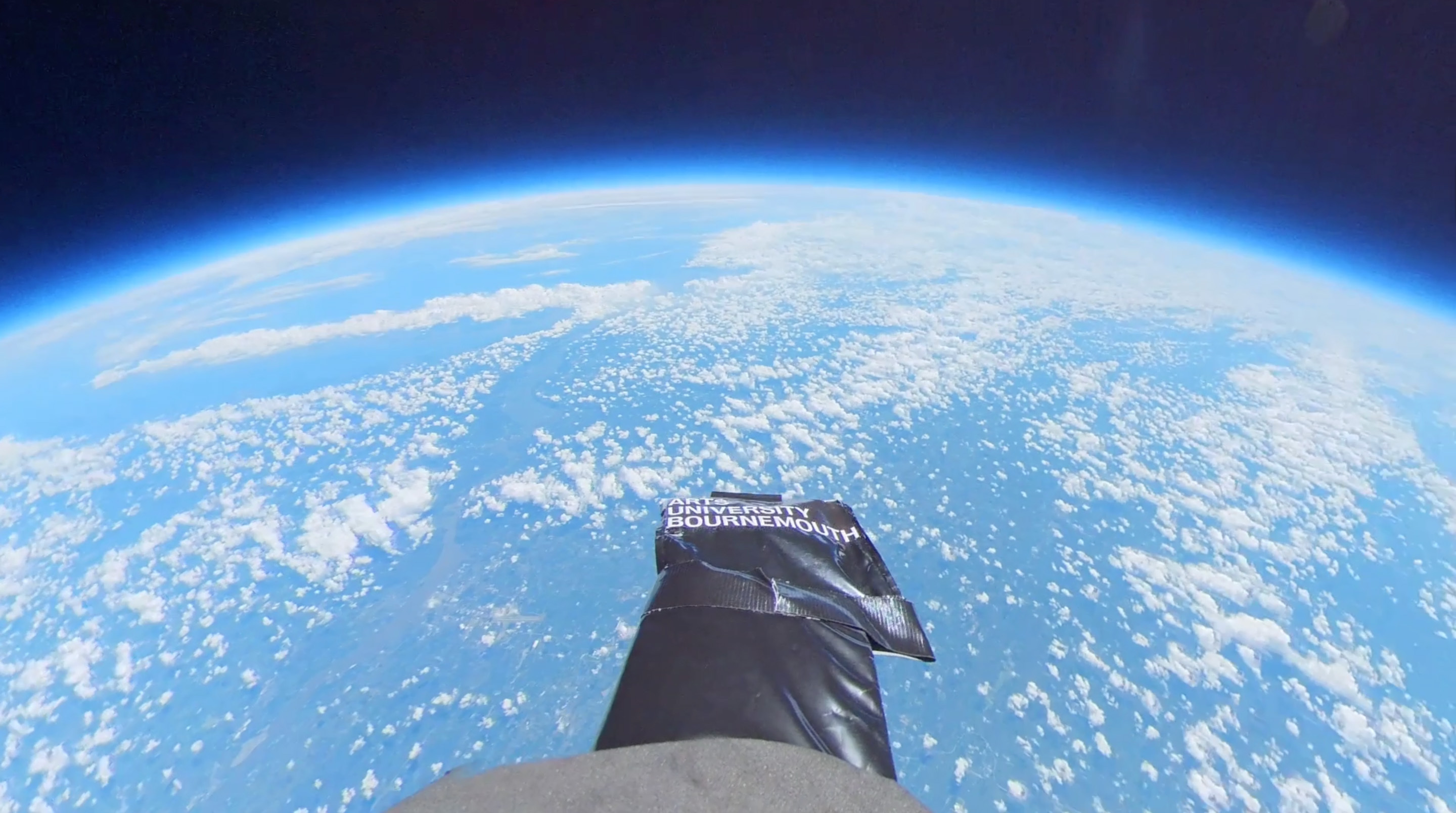 Black boot in foreground against curved Earth horizon showing blue and white cloud patterns, dark space above with blue atmospheric glow.