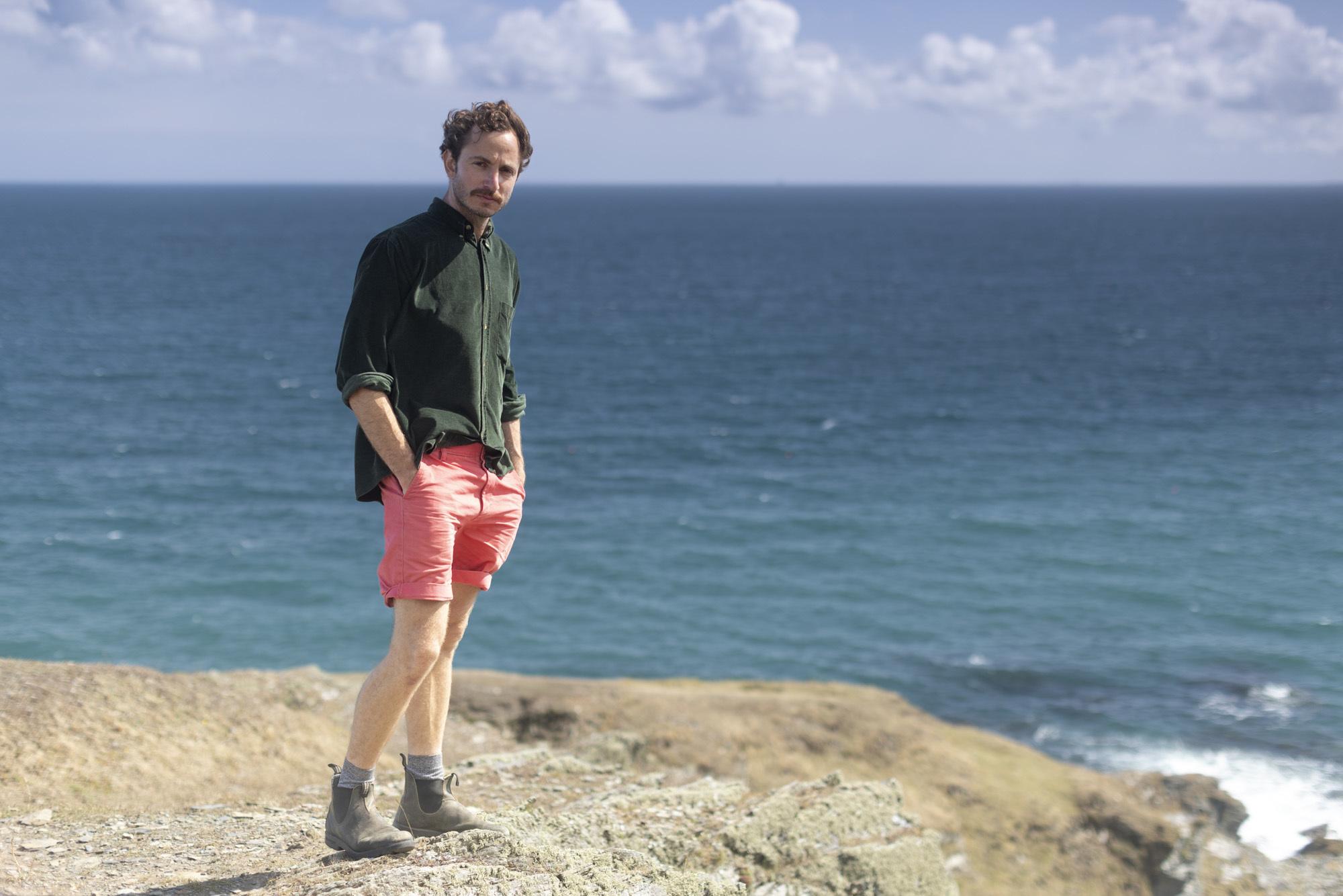 Tom Massey wearing a green top and pink shorts stood on a tall cliff, blue sea and sky in the background.