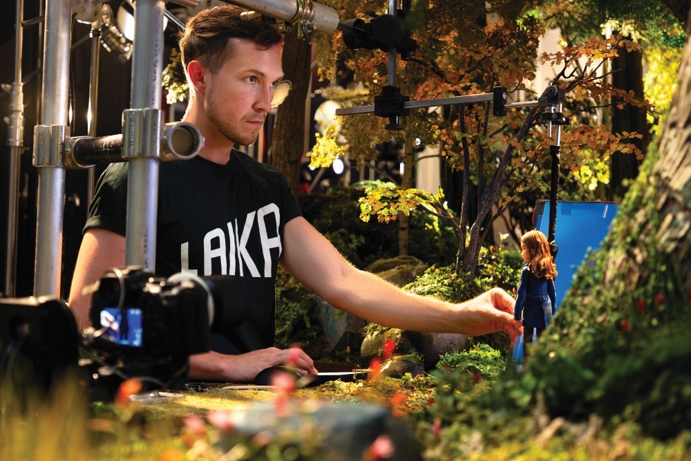 A man in a black LAKA T-shirt adjusts a miniature outdoor set, with a small figure and filming equipment visible around him.