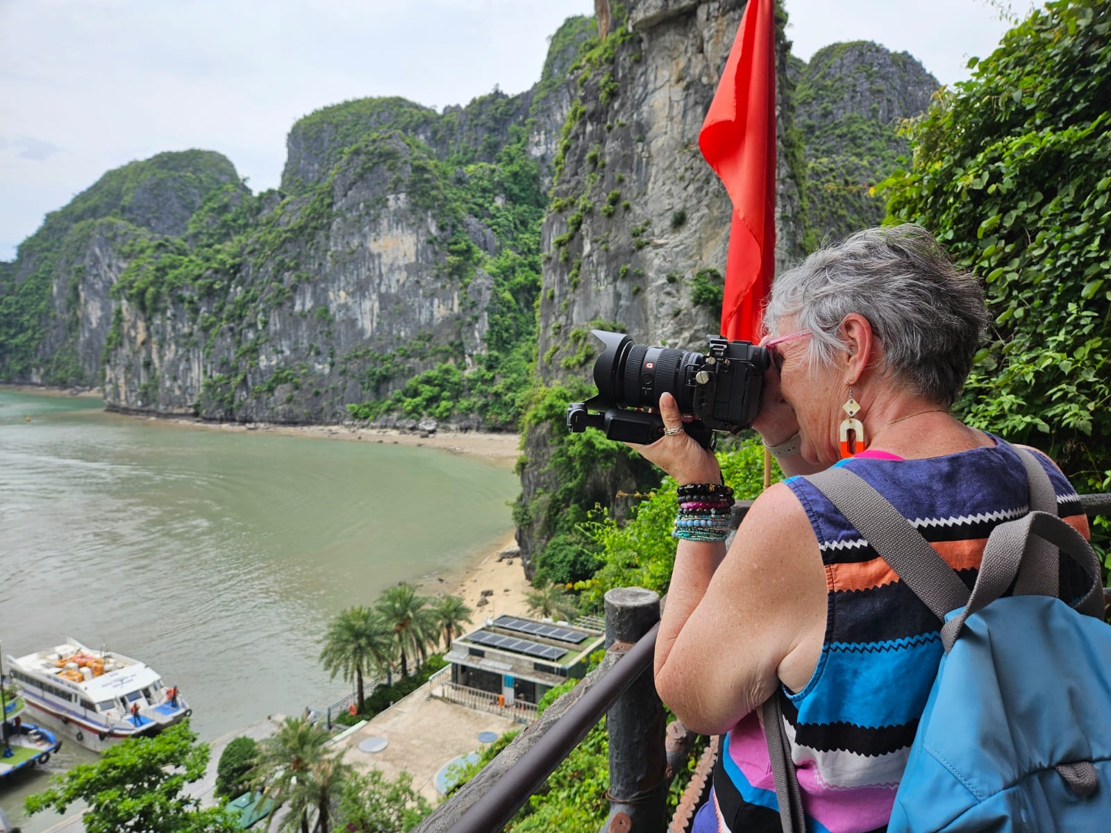 A woman with a blue backpack photographs a limestone bay from a viewpoint, with boats and a sandy beach below.