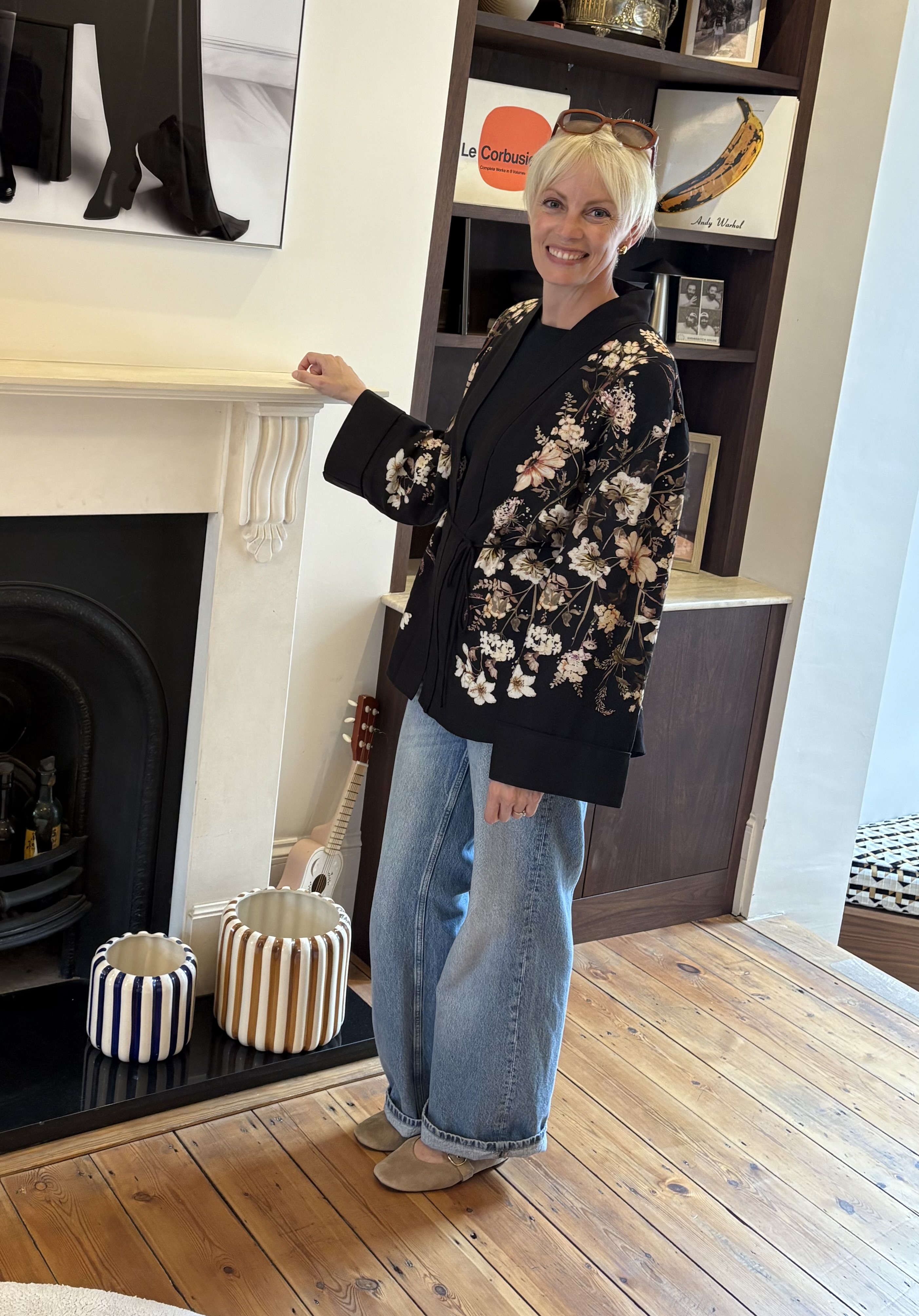 A woman wearing a black floral cardigan and wide-leg jeans stands beside a white fireplace, with striped ceramic stools and shelves behind her.