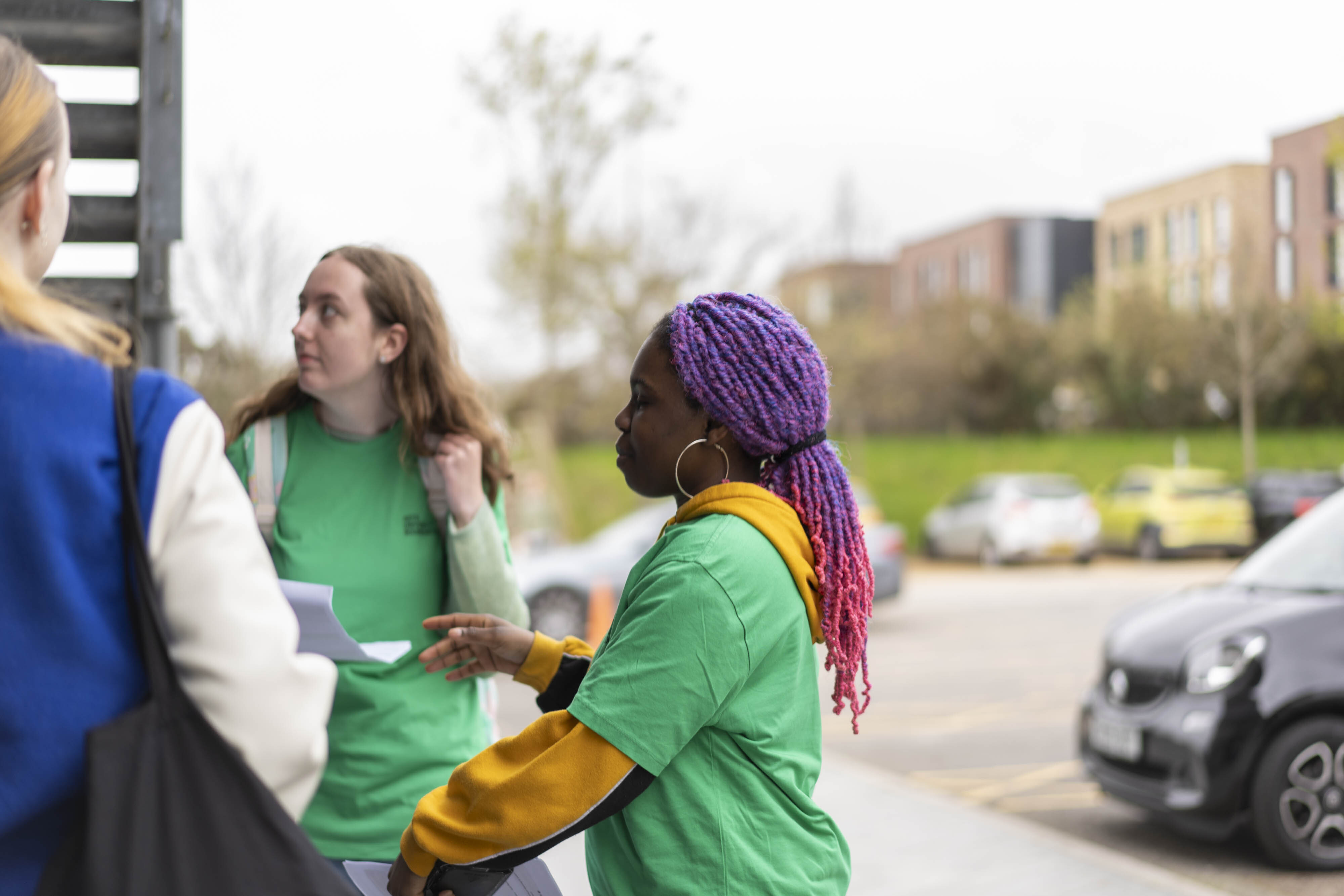 Three women in conversation outdoors, two wearing green tops, one in blue jacket. Woman on right has purple braided hair.