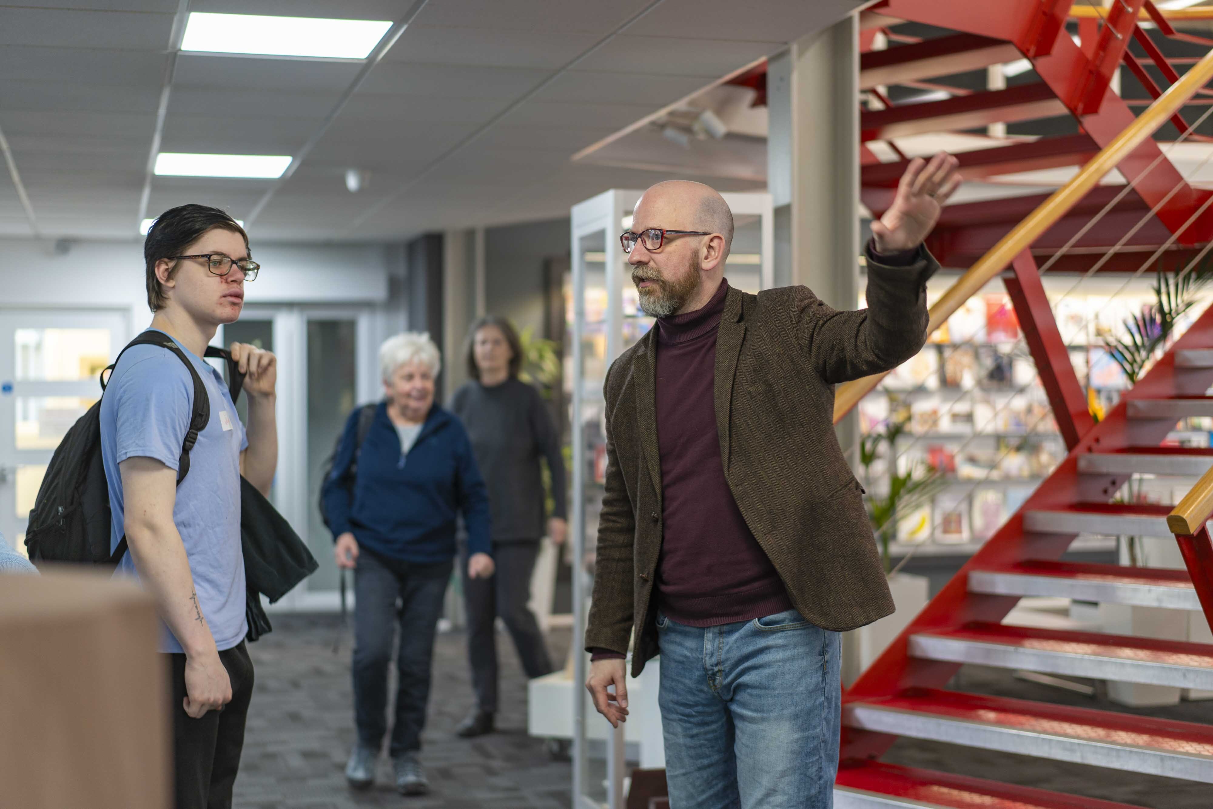 Bald man in glasses and brown jacket gestures upward whilst speaking to younger man with rucksack in modern building interior.
