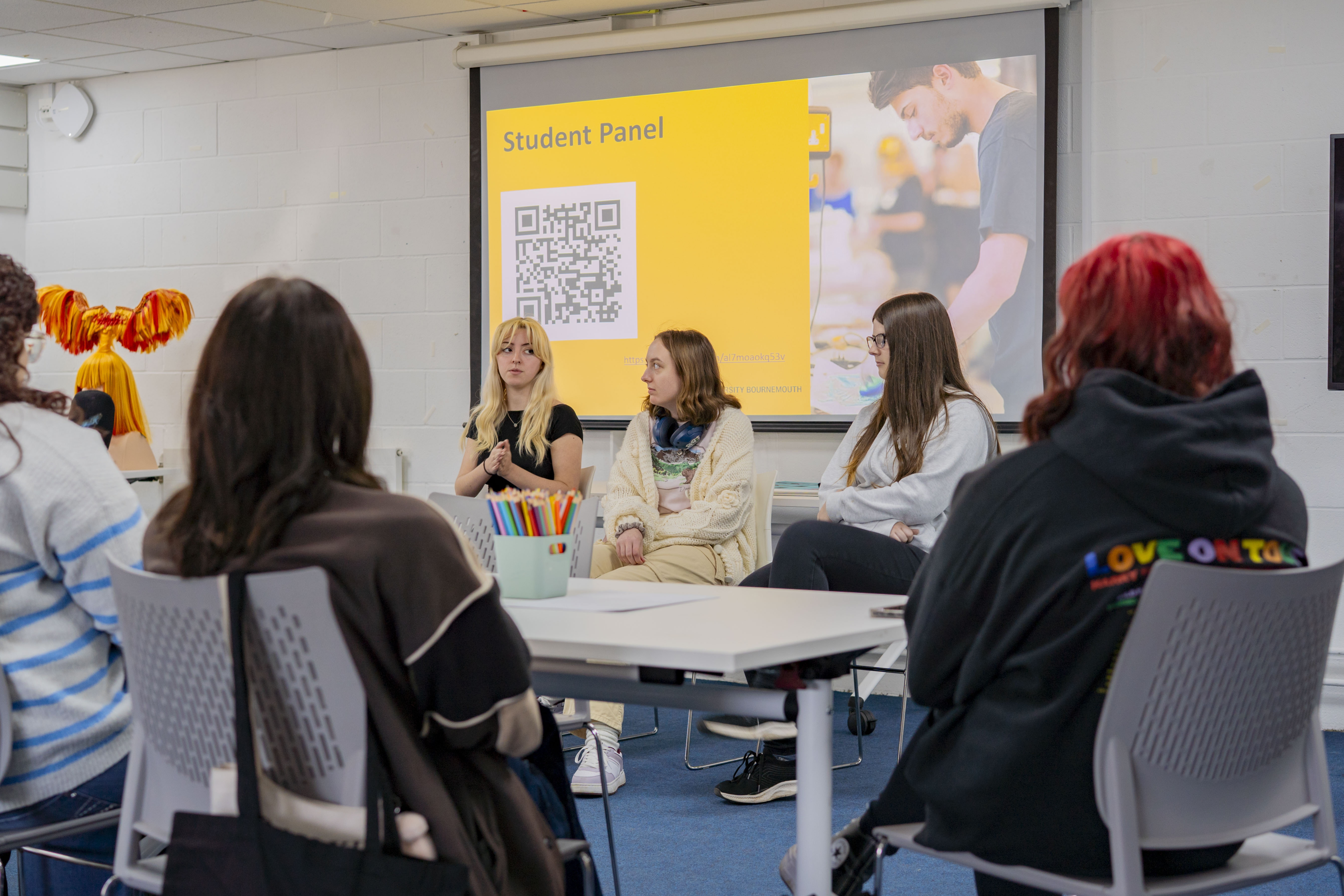 Students seated at white tables in classroom facing yellow projection screen displaying "Student Panel" text and QR code.
