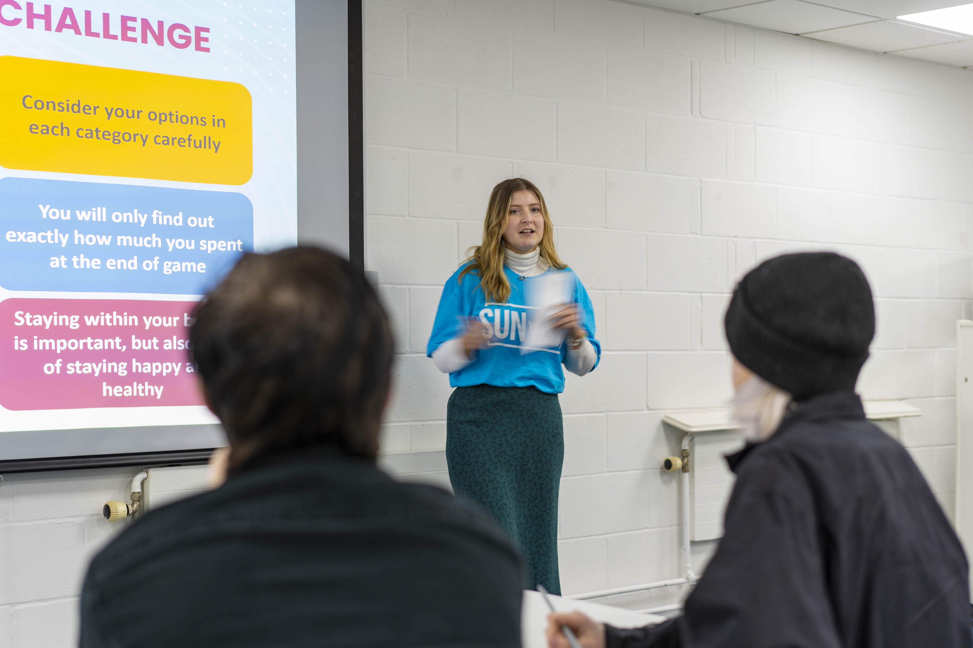 Woman in blue jumper presenting to audience in classroom with colourful projected slide about challenges visible on wall.