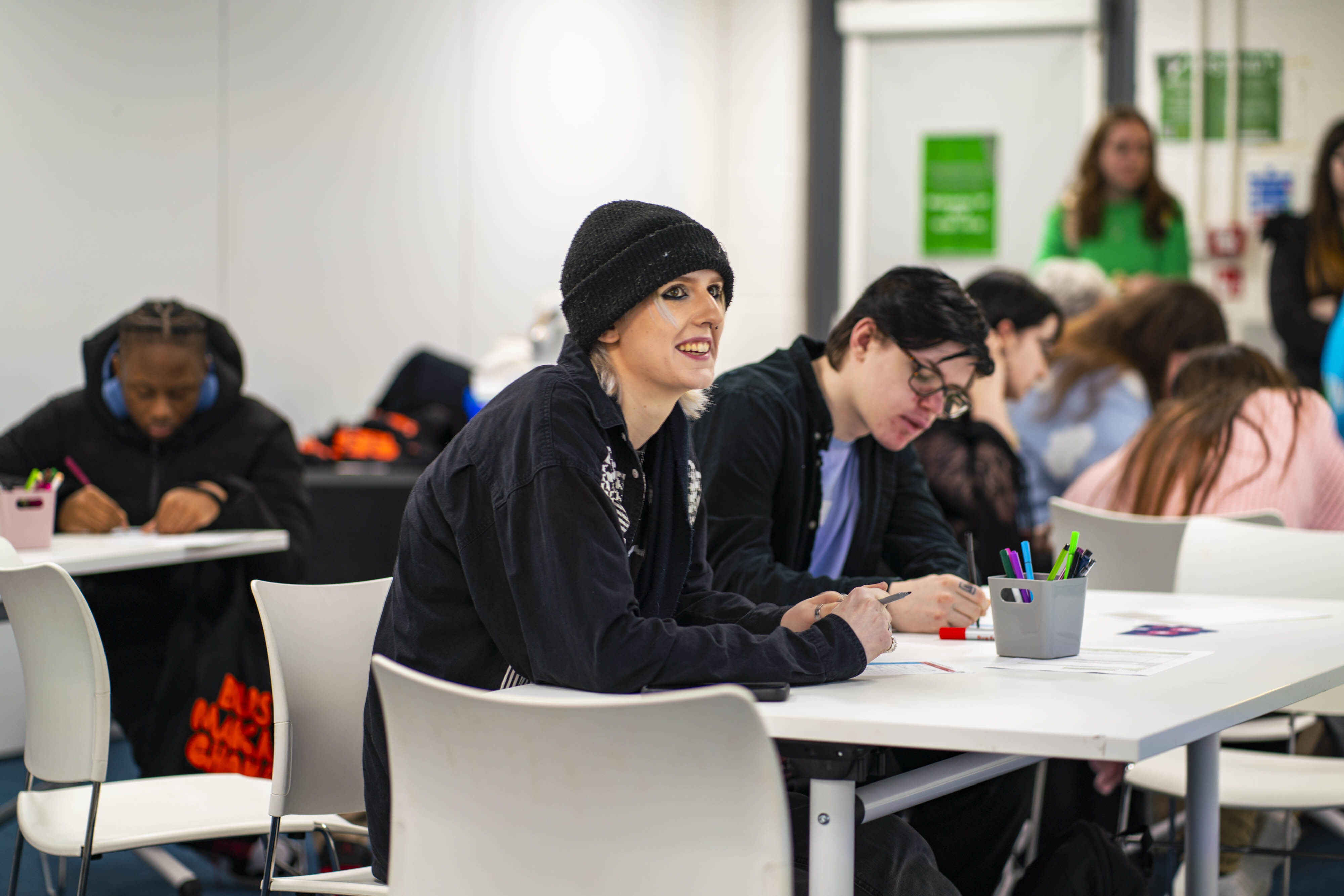 Students seated at white tables in classroom, woman in black hoodie and beanie in foreground, teacher in green top visible in background.