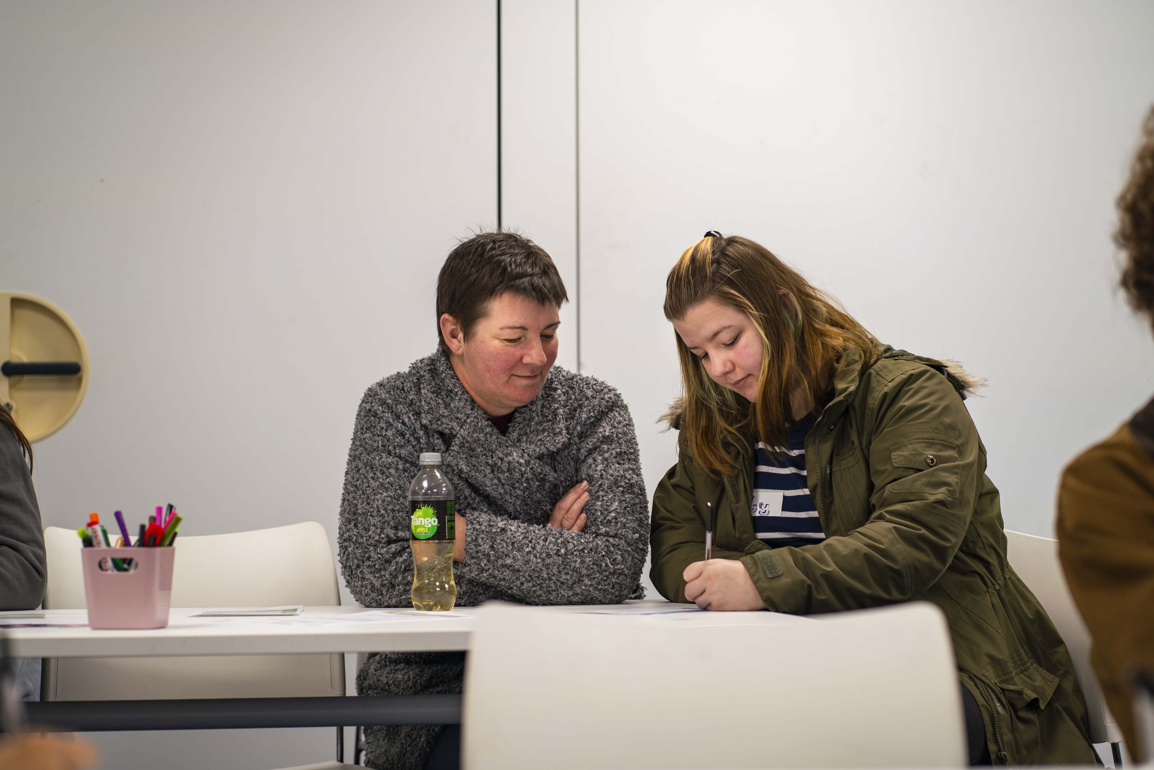 Two women sitting at white table in modern office. One wears grey jumper, other in olive green jacket over striped top. White chairs visible.