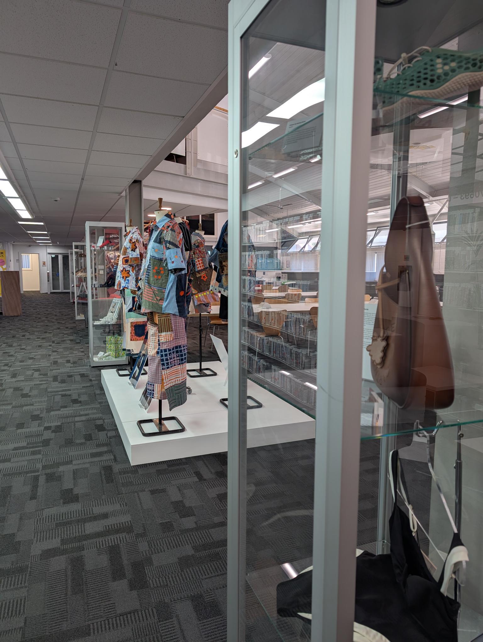 Glass display case in library containing colourful textile garments on stands, with bookshelves visible in background.