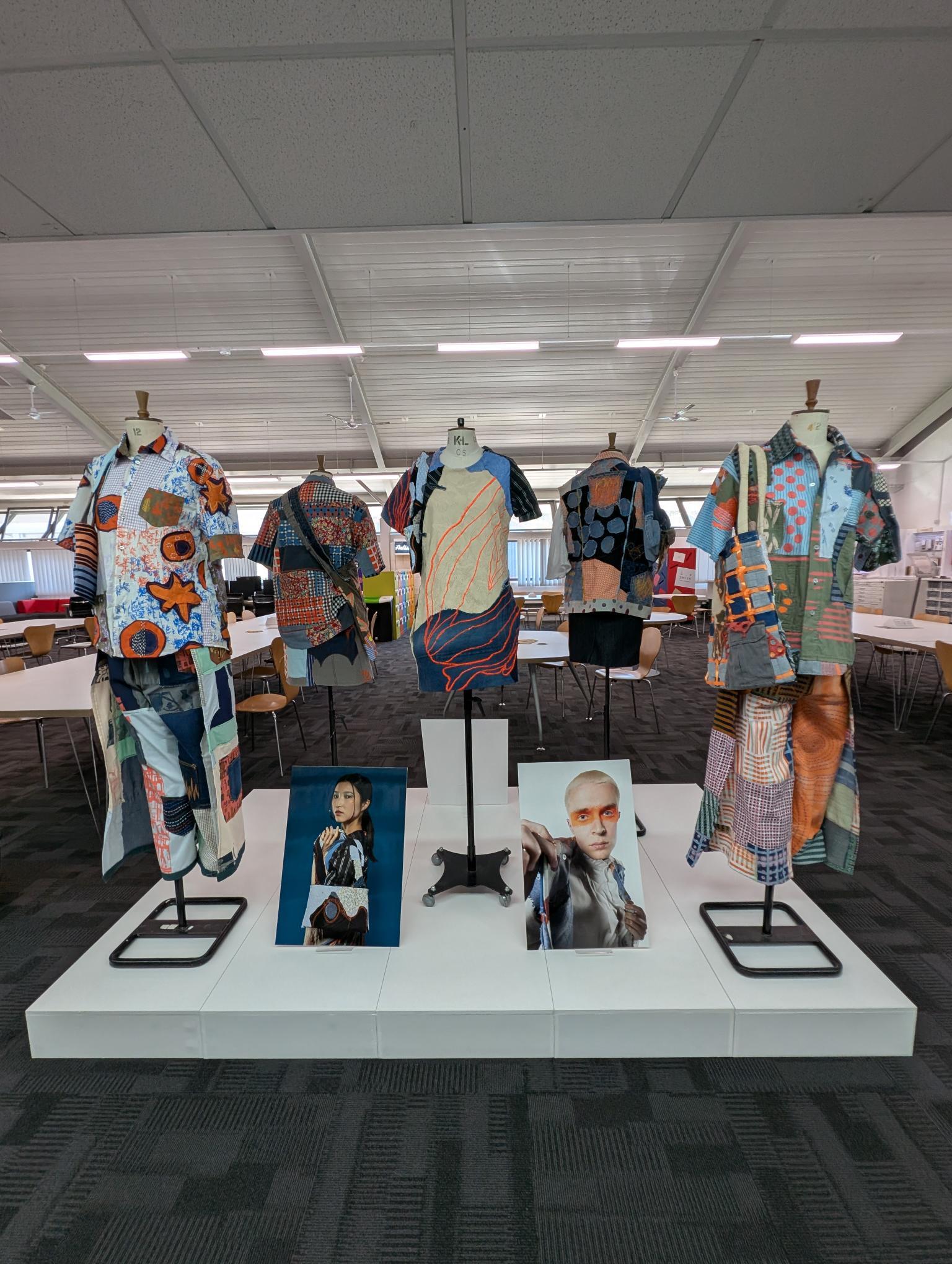Fashion exhibition display with colourful patterned garments on mannequins in white warehouse space with suspended ceiling lights.