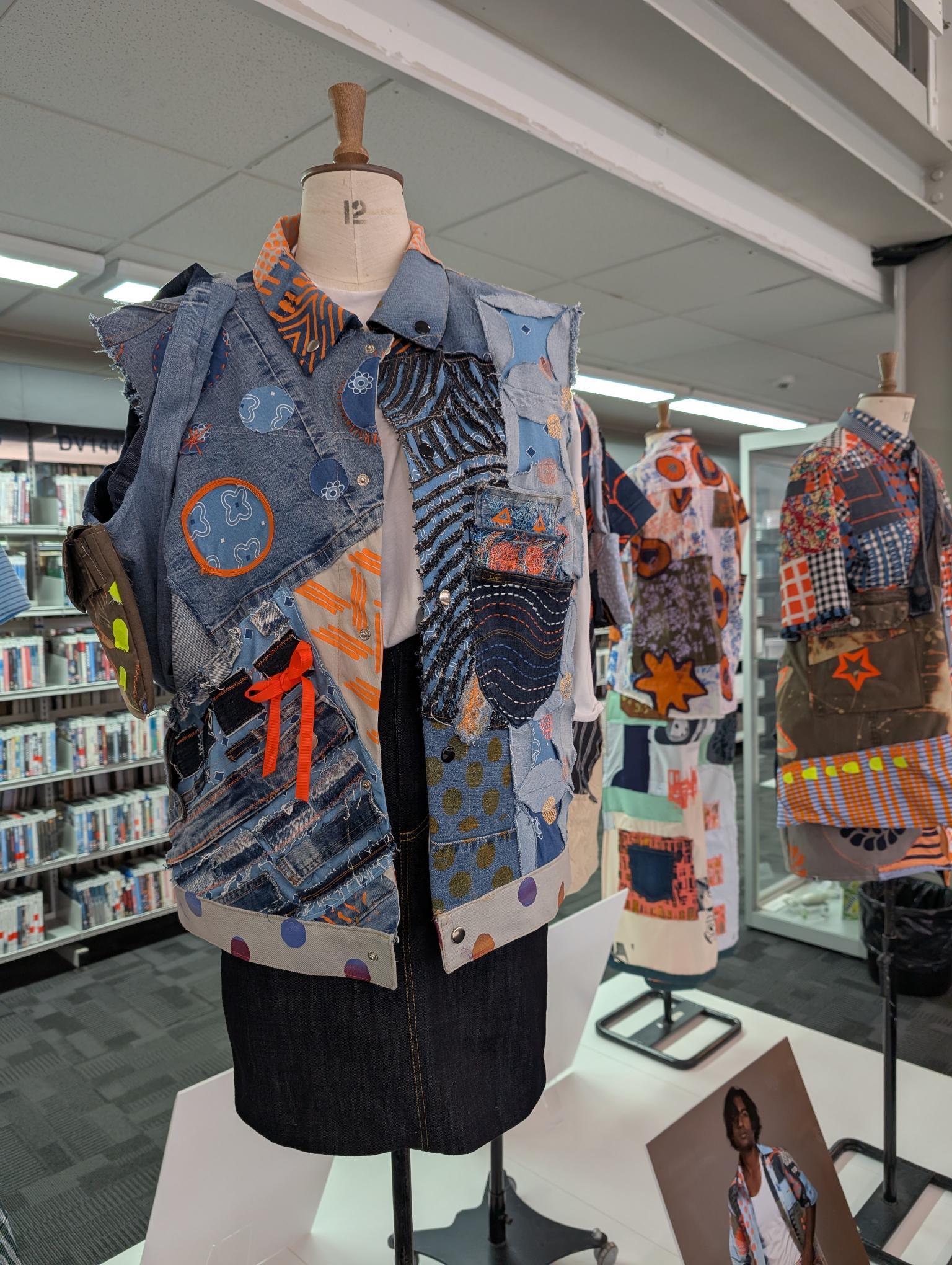 Dressmaker's mannequin wearing patchwork jacket with blue denim, orange geometric patterns, and mixed fabric pieces in library setting.