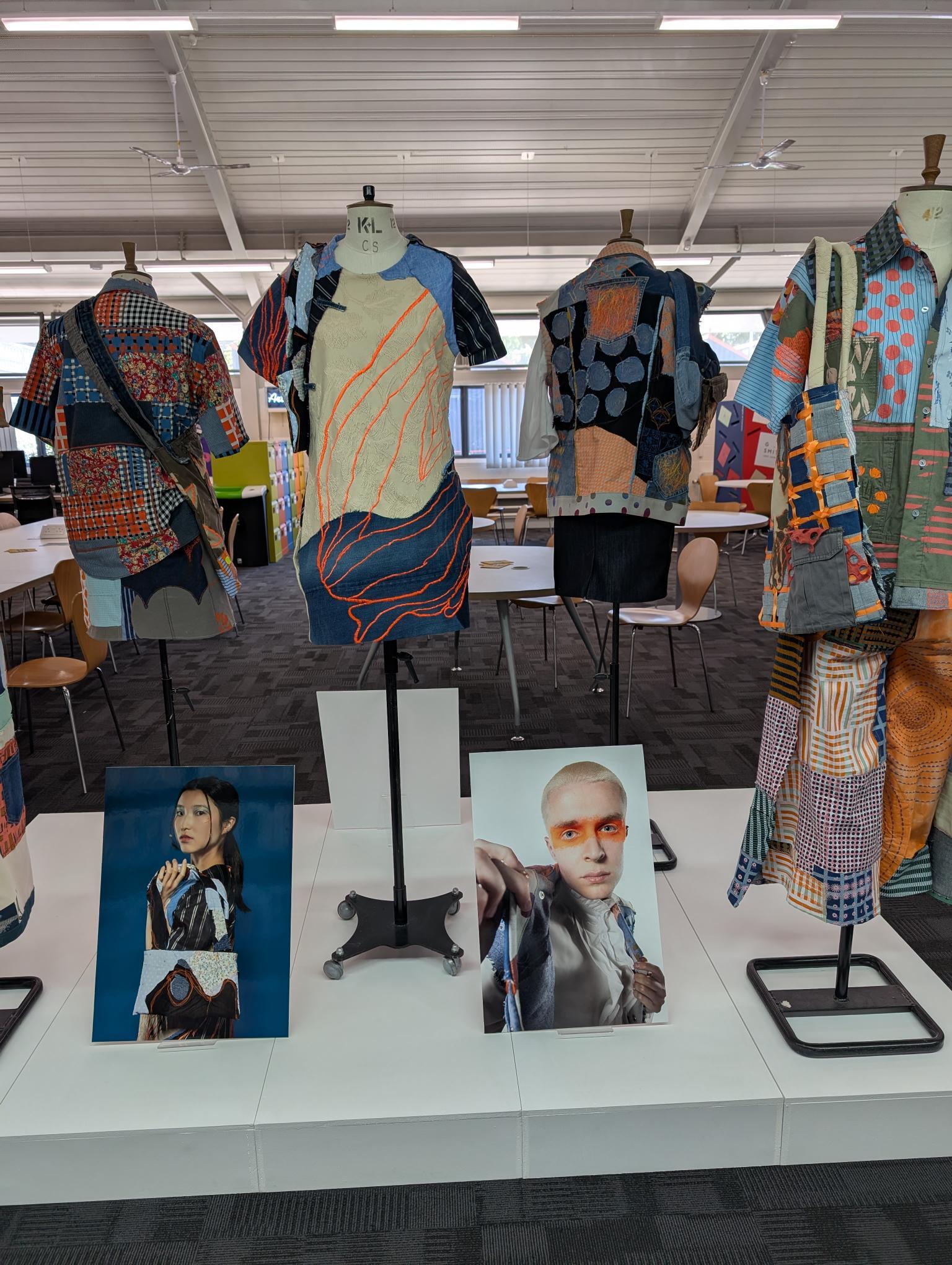 Fashion design studio with dress forms displaying colourful patterned garments and fabric samples. Portrait photographs on white table below.