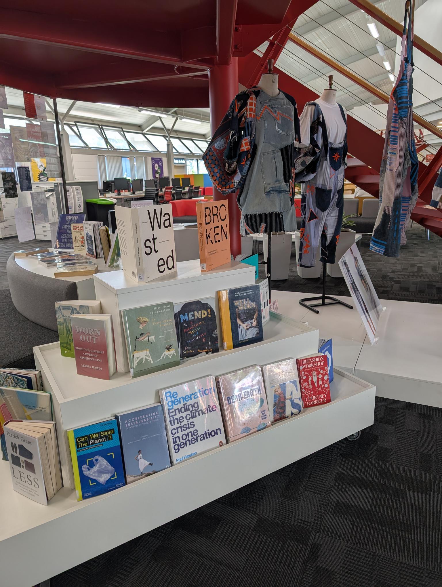 Library display with white tiered shelving showing books, clothing on mannequin, and cardboard signs reading "Wasted" and "Broken".