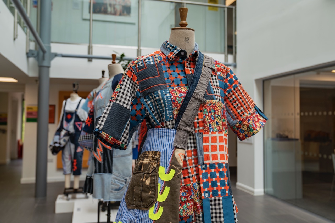Showcase exhibition for 'Trash or Treasure' showing a collection of clothes lined up on a display stand in University House, each one having a patchwork appearance with blue, orange and pink textiles.