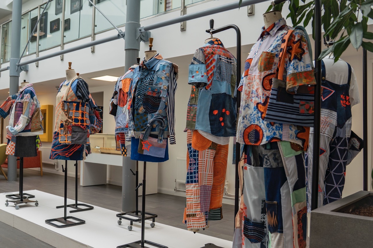 Showcase exhibition for 'Trash or Treasure' showing a collection of clothes lined up on a display stand in University House, each one having a patchwork appearance with blue, orange and pink textiles.
