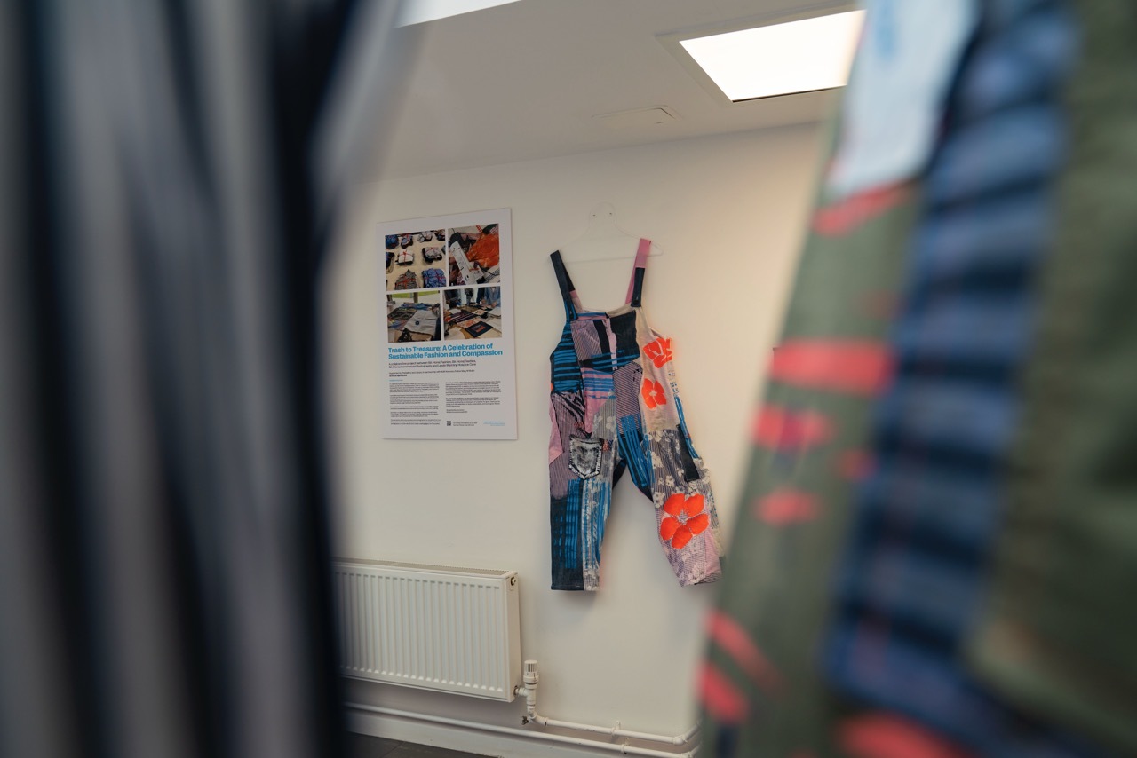 Part of the Trash to Treasure exhibition, showing colourful dungarees and text on a white wall in-between blurred clothing.