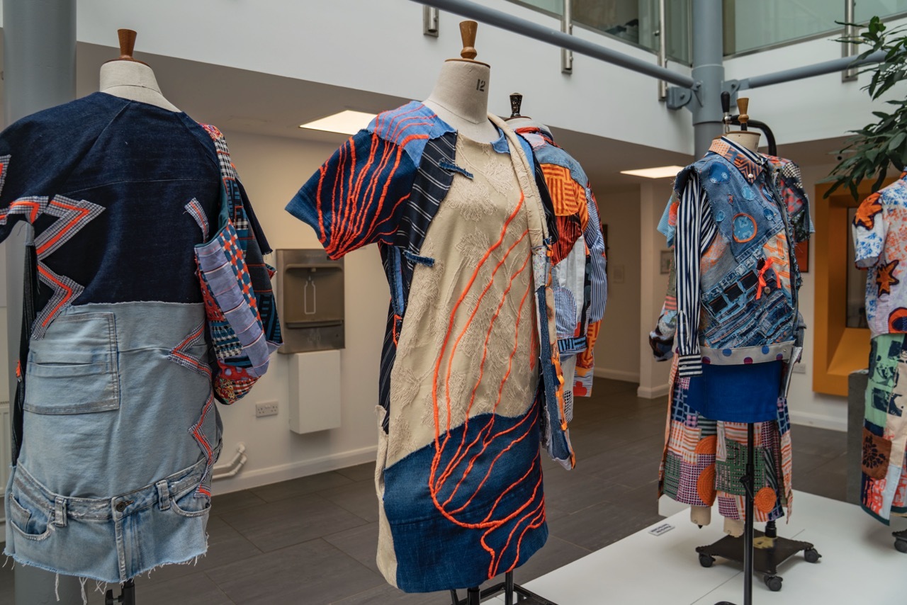 Showcase exhibition for 'Trash or Treasure' showing a collection of clothes lined up on a display stand in University House, each one having a patchwork appearance with blue, orange and pink textiles.