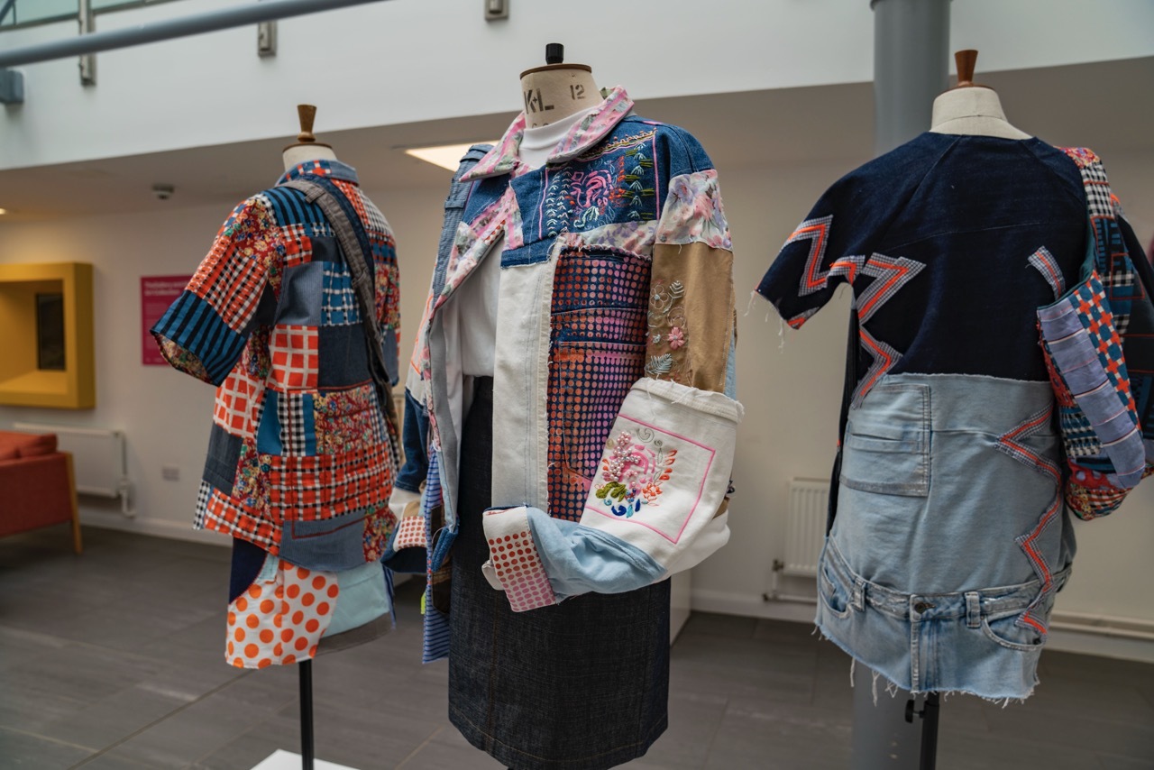Showcase exhibition for 'Trash or Treasure' showing a collection of clothes lined up on a display stand in University House, each one having a patchwork appearance with blue, orange and pink textiles.
