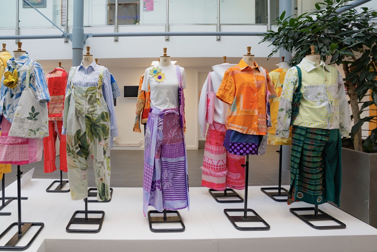 Colourful garments displayed on mannequins in modern gallery space with white floors and walls, featuring bright fabrics in orange, purple, green, and mixed patterns