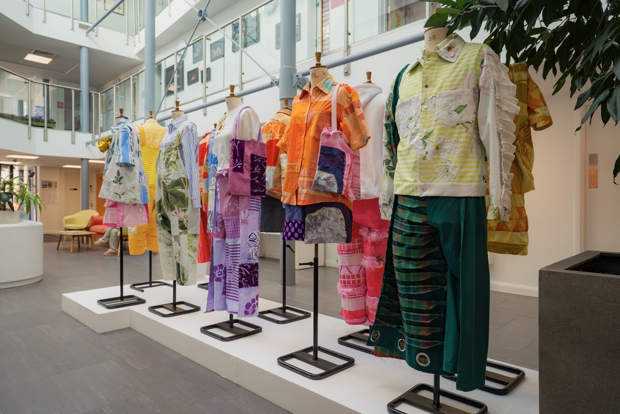 Fashion exhibition with colourful garments on black stands: orange, purple, yellow, green, and patterned pieces in modern gallery space