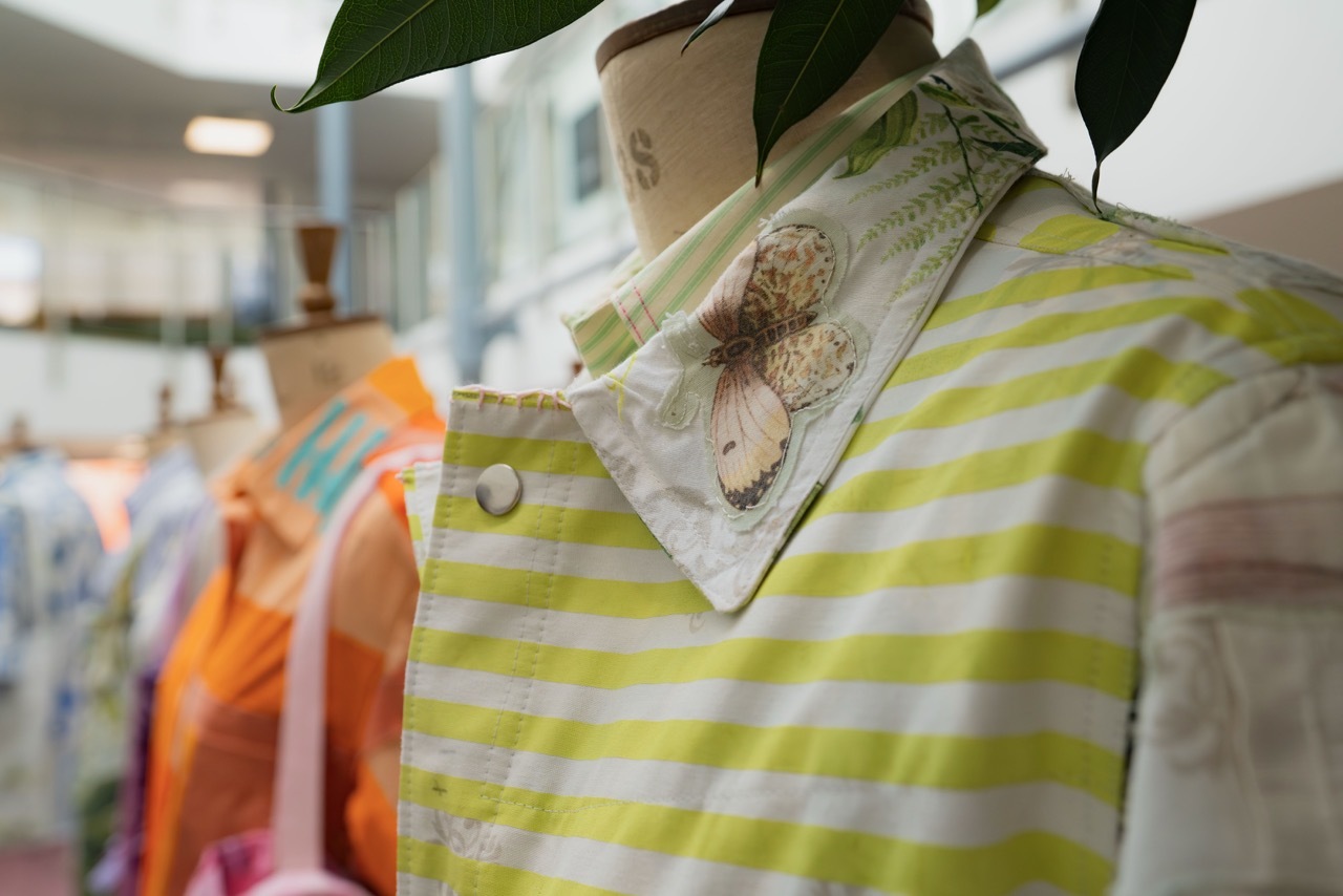 Green and white striped garment on mannequin with decorative patch, green leaves above, orange clothing visible in background