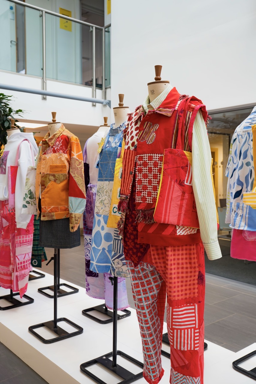 Clothing display with mannequins wearing patchwork garments in red, orange, blue and white patterns on metal stands in gallery space