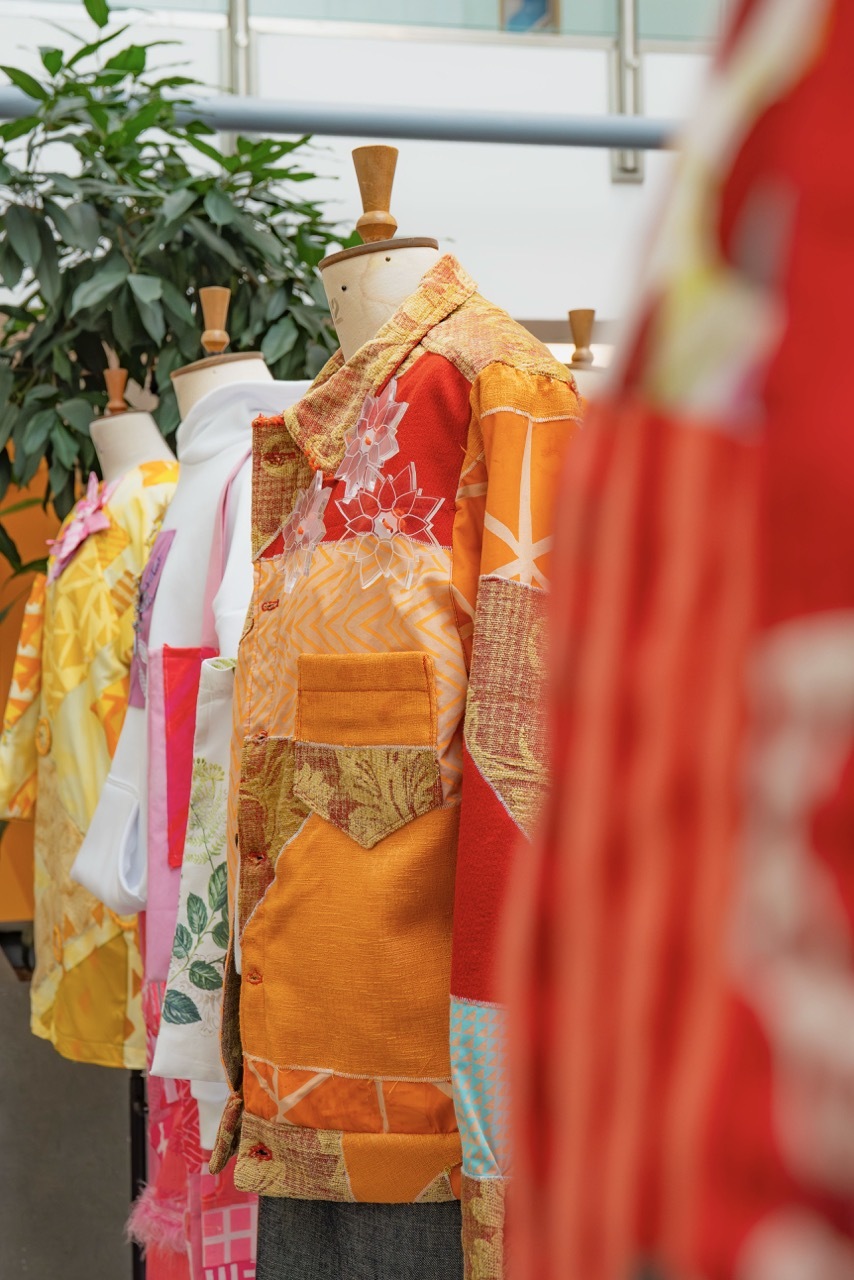 Colourful kimono displayed on wooden dress form with orange, red and yellow patchwork patterns, green plant visible in background