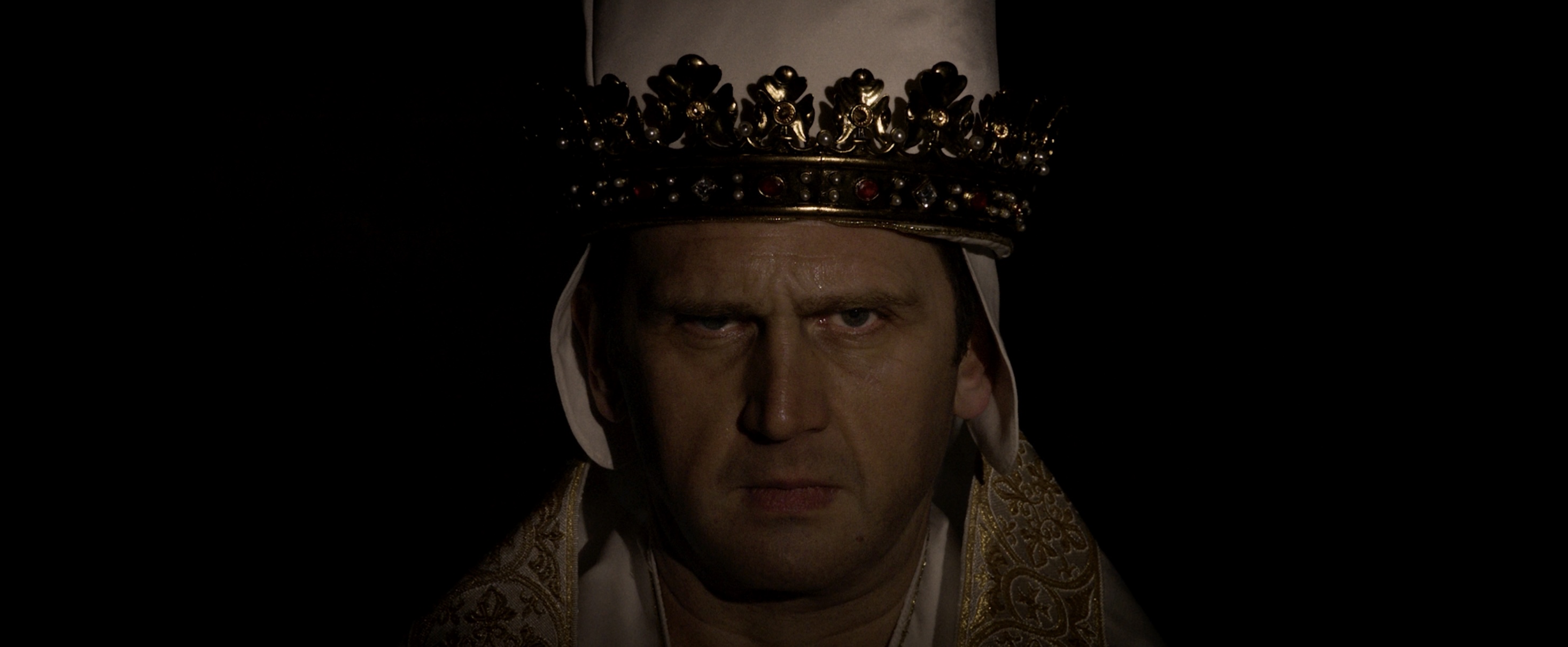 A man wearing a white papal robe adorned with gold embroidery, topped with an ornate gold crown. The background is dark, creating a dramatic contrast with the figure's features and clothing.