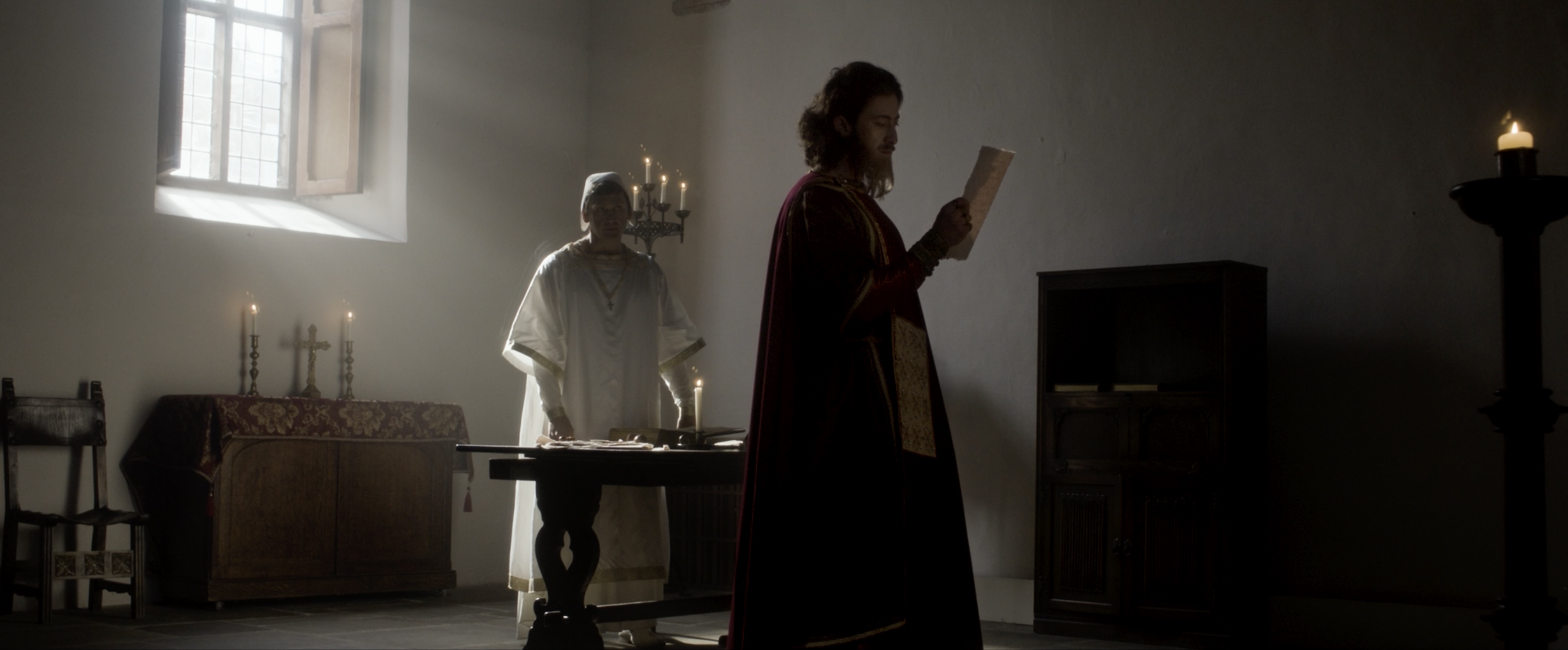 A person in a long maroon robe stands in a dimly lit room, reading from a parchment. A statue of a man in white robes is positioned behind them, next to a wooden table with candles on it, set against a wall with a window.