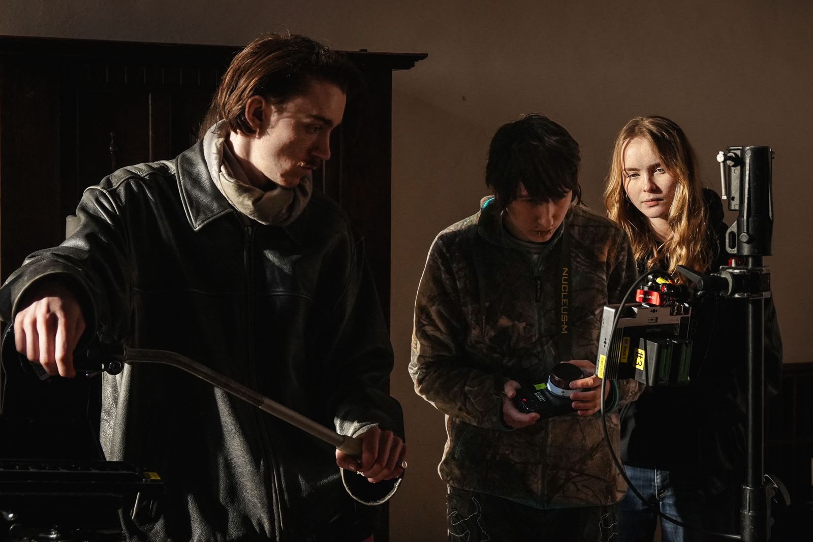 Three individuals are working together on a film set. One person, wearing a black jacket, is adjusting equipment, while another, dressed in a camouflage hoodie, is holding a camera. The third person, a woman in a light-coloured top, observes both of them.
