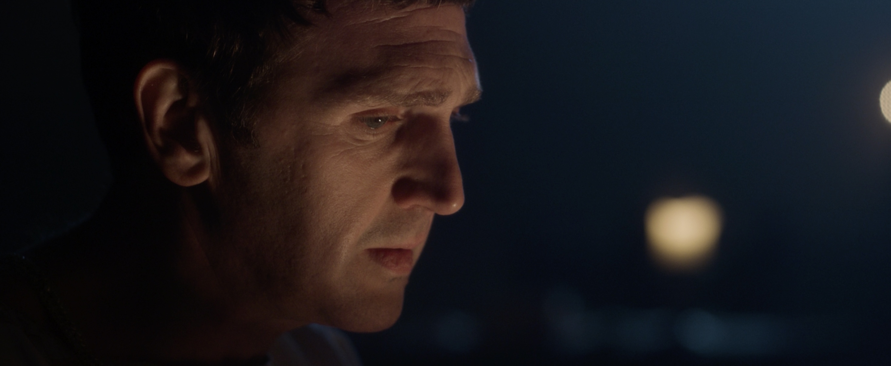A close-up of a man with short hair, facing slightly left. The scene is dimly lit, with soft light illuminating his profile against a dark background. There are faint light sources creating a bokeh effect in the background.