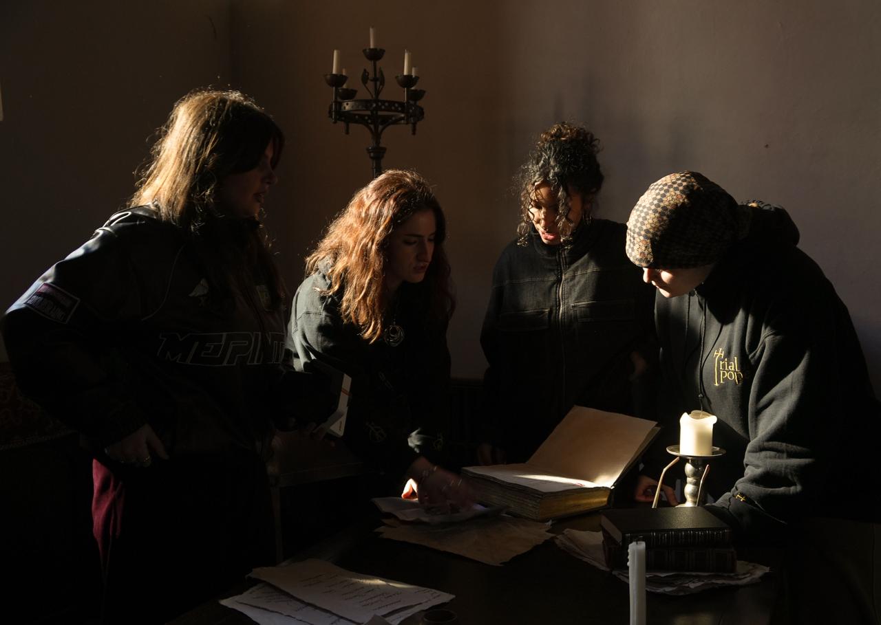 Four people stand around a table in a dimly lit room. They are reviewing documents and a large open book, with a candle and a candelabrum visible on the table, casting soft light.
